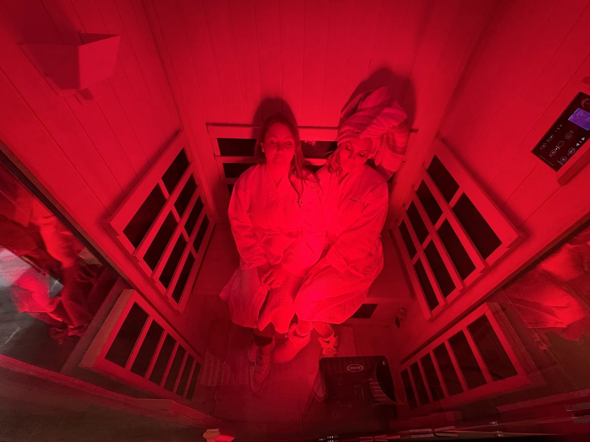 Two women lying back-to-back in a sauna bath with red lighting, wearing robes, with wooden walls and a control panel visible.