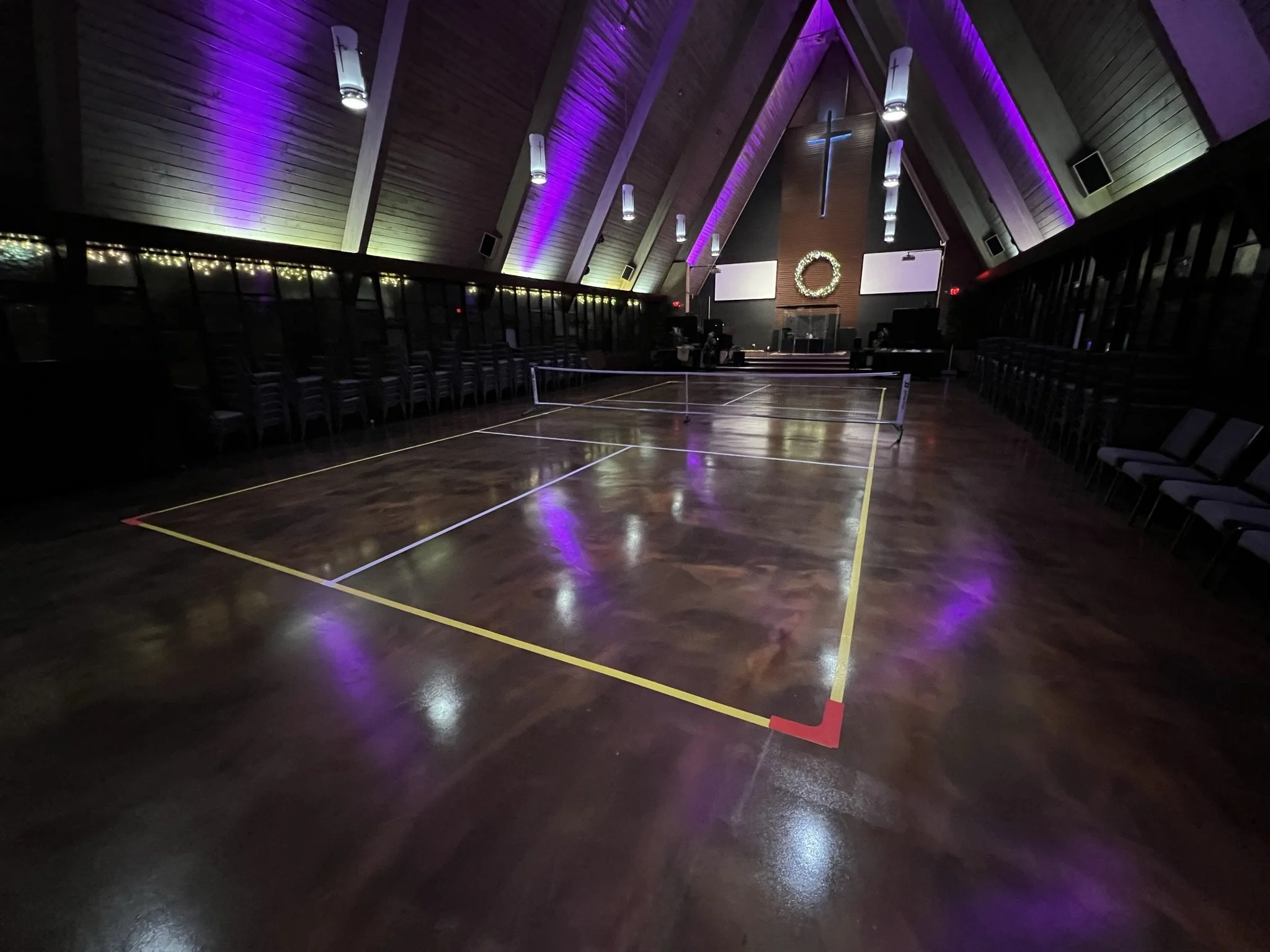An empty church or chapel with a wooden ceiling and purple and white lighting. There is a large cross on the wall behind the stage and a wreath hanging on the wall. The floor is a polished wooden floor with a badminton net set up in the middle.