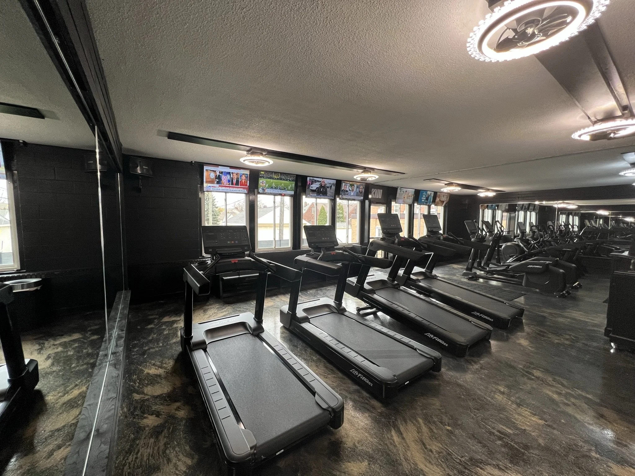 Row of black treadmills inside a gym with multiple monitors on the wall displaying news and sports.