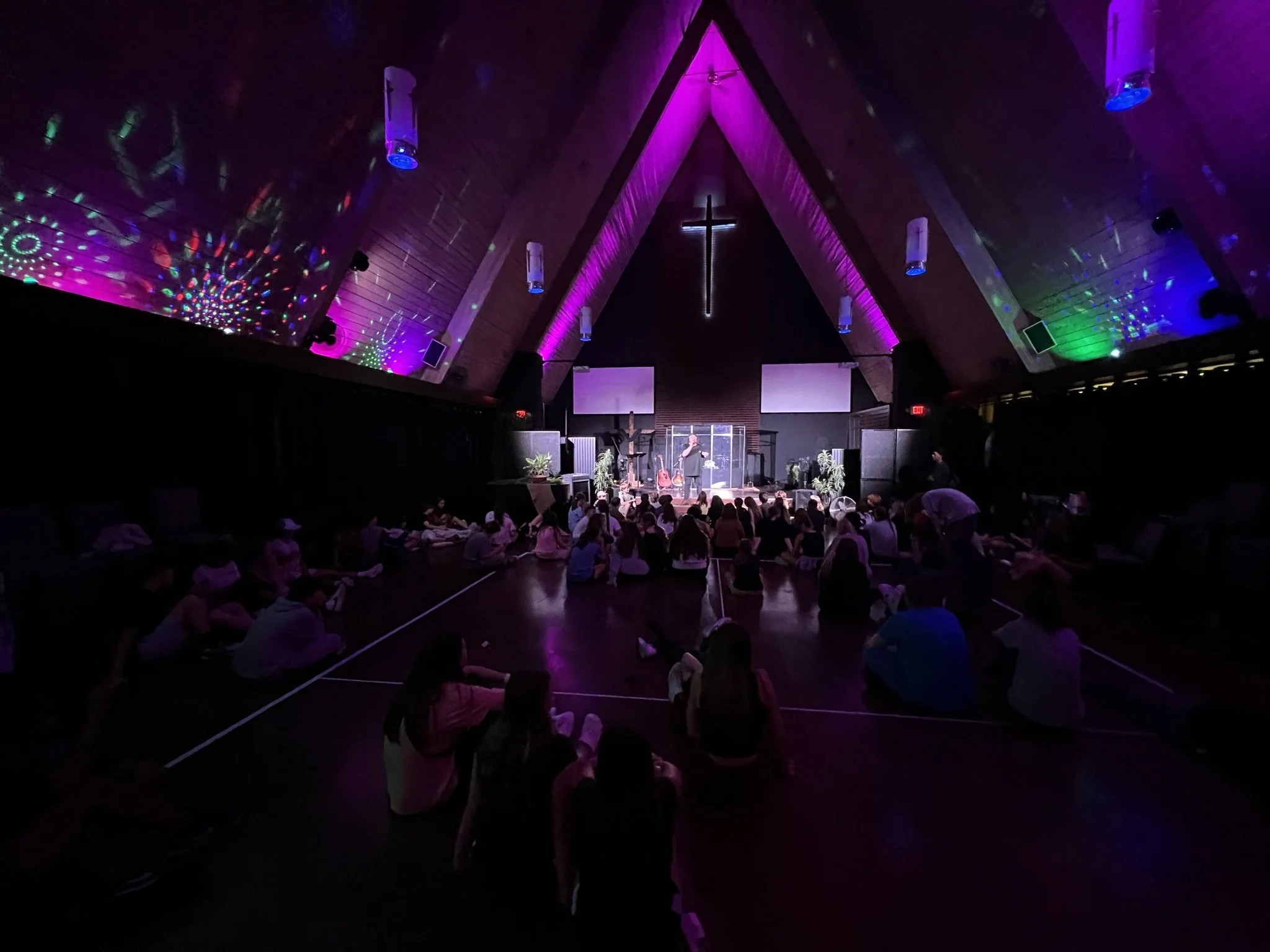 Inside a church with wooden A-frame ceiling, colorful lighting, a cross at the front, and people seated on the floor.