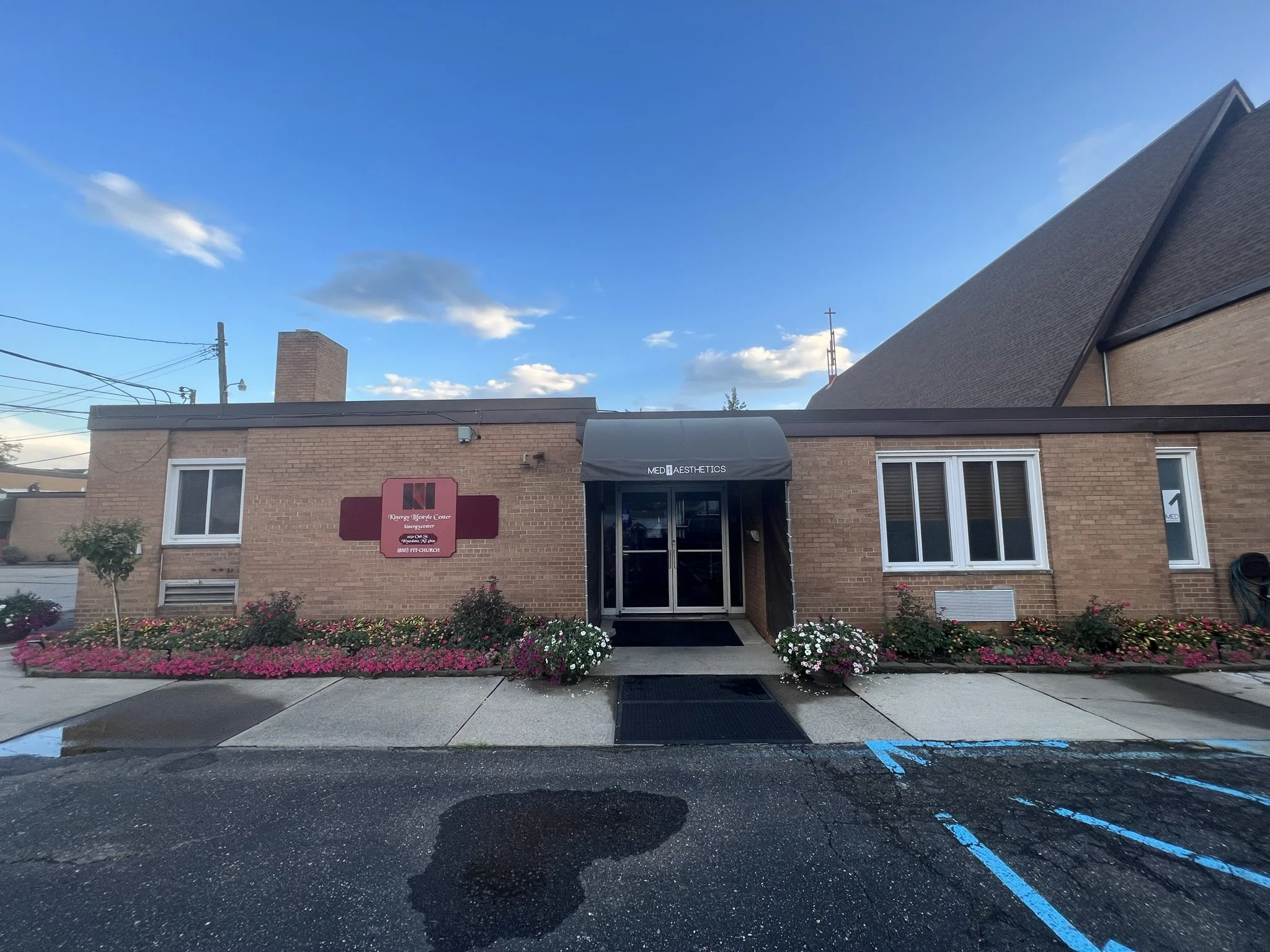 Front view of a brick building with a black awning labeled 'MED AESTHETICS' and red sign with white text, surrounded by pink and white flowers, with a blue sky and clouds above. There are parking spaces in the foreground and power lines overhead.