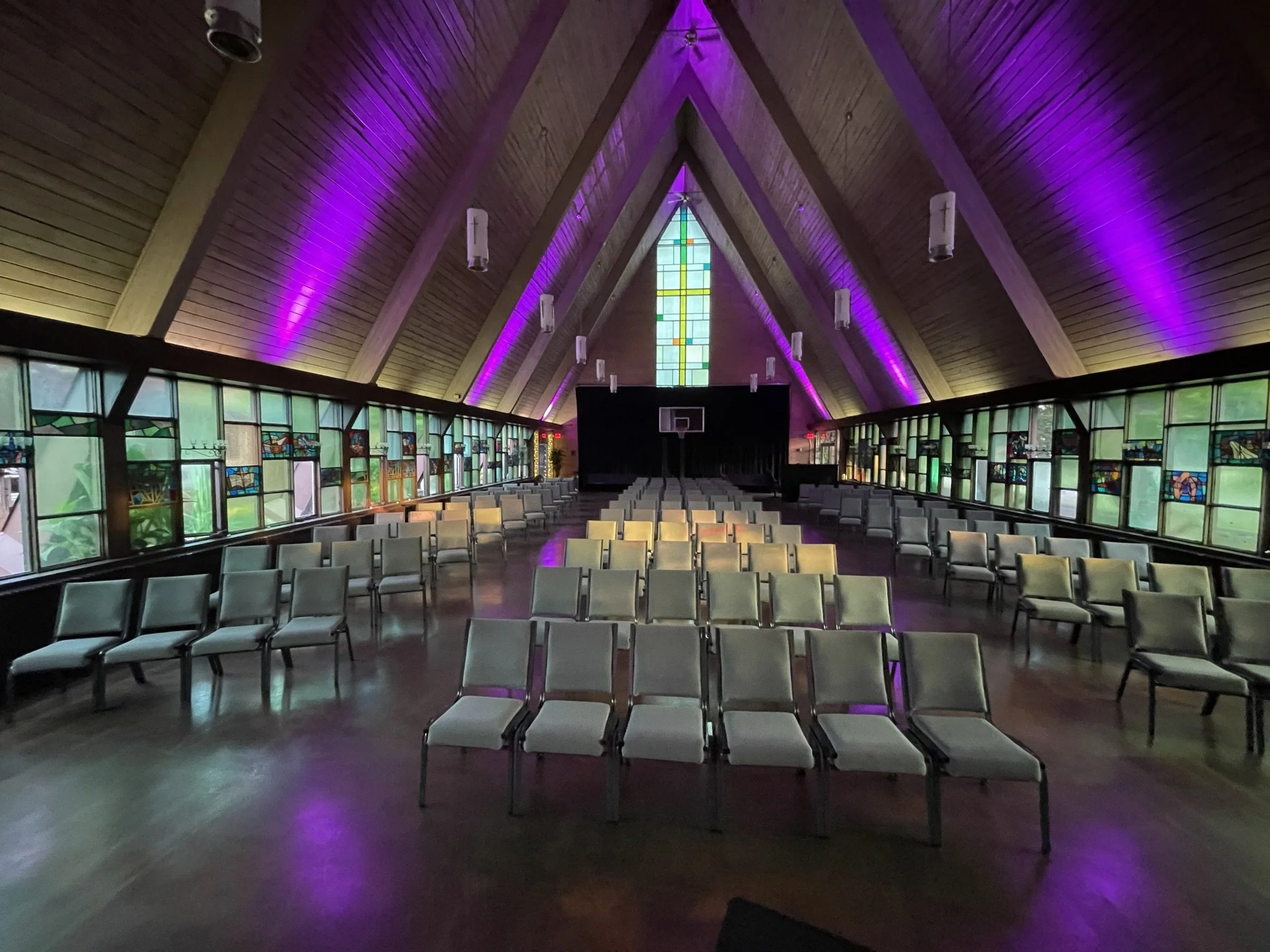 Interior of a church sanctuary with rows of white chairs, stained glass windows, a wooden ceiling with purple lighting, and a basketball hoop at the front.