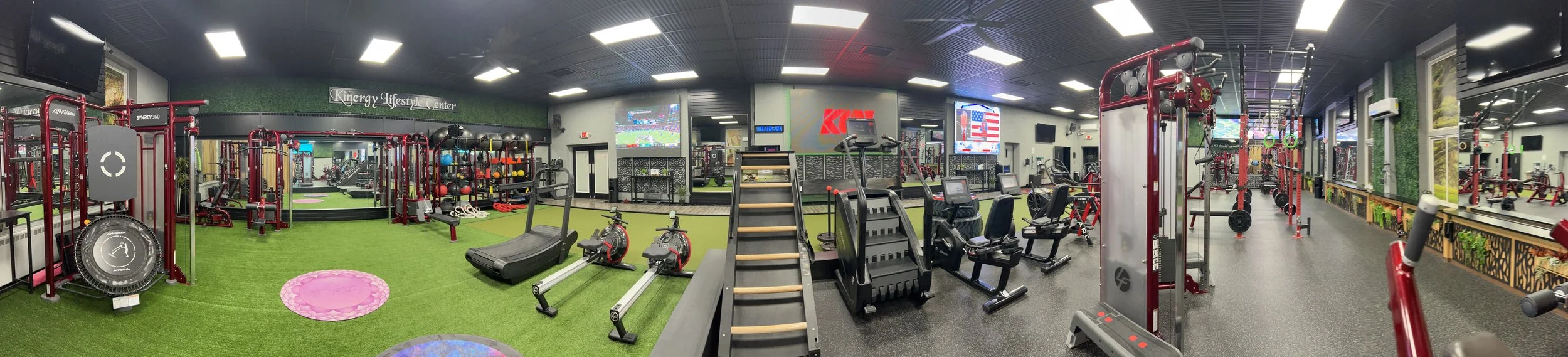 A panoramic view of a fitness center with various exercise equipment, including treadmills, stationary bikes, free weights, and resistance machines, with a large television screens on the wall and a sign that reads 'Kenergy Lifestyle Center'.