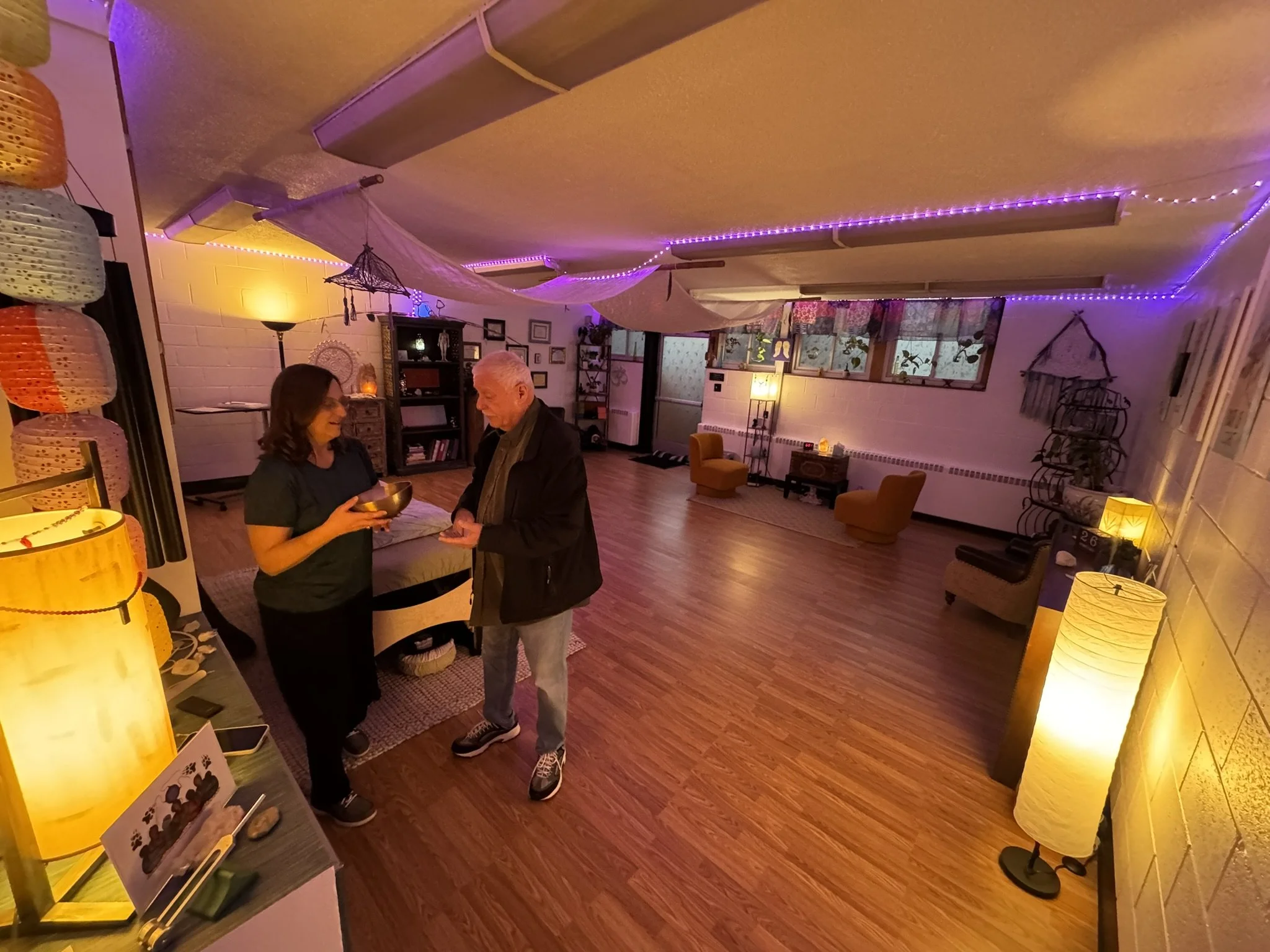 Two people, a woman and a man, stand inside a warmly lit room with colorful lighting and decorations. The woman holds a bowl and appears to be explaining something to the man. The room has cozy furniture, hanging lights, and decorative items on shelv