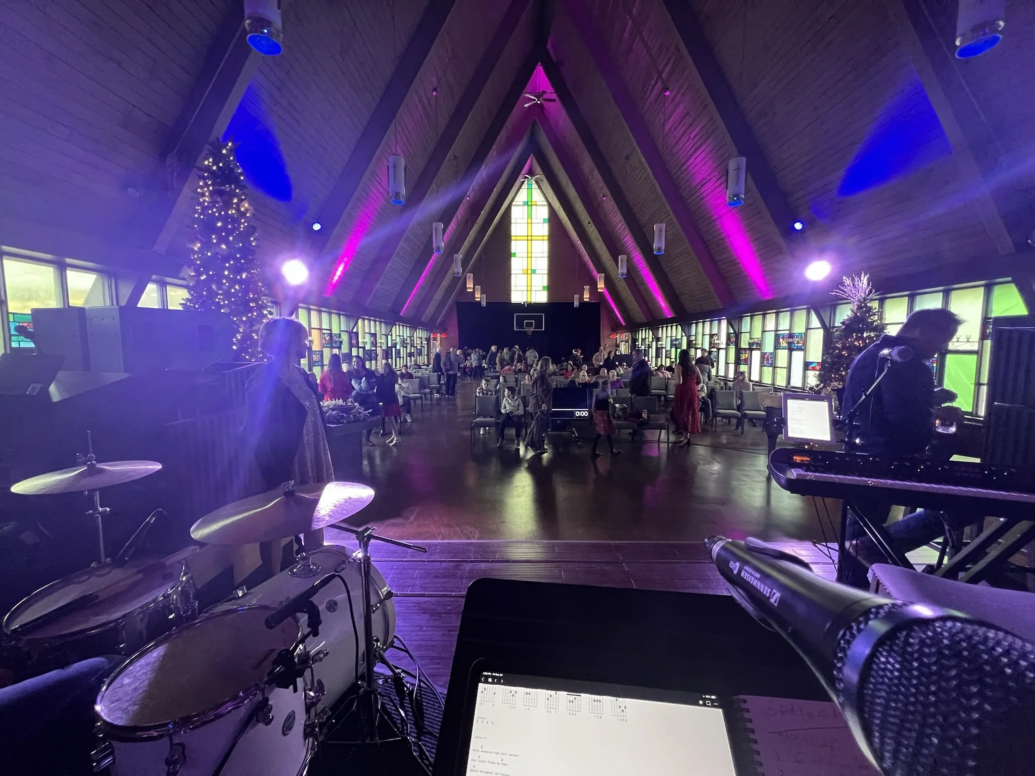Interior of a church or chapel decorated with Christmas trees and colorful lighting, with people gathered inside, and a person playing electronic keyboard, along with a drummer and a singer.