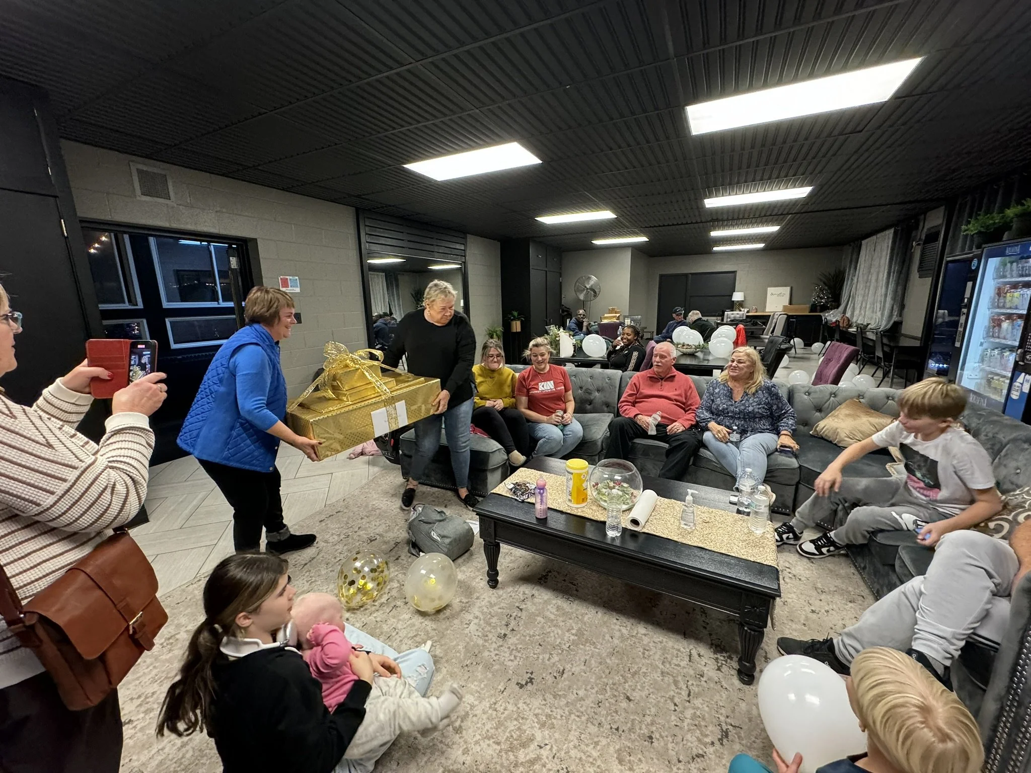 People gathered in a living room celebrating, with balloons and a large wrapped gift on a table. Two women are handing the gift to a woman in a blue jacket, who is smiling. Others sit on sofas, some taking photos.