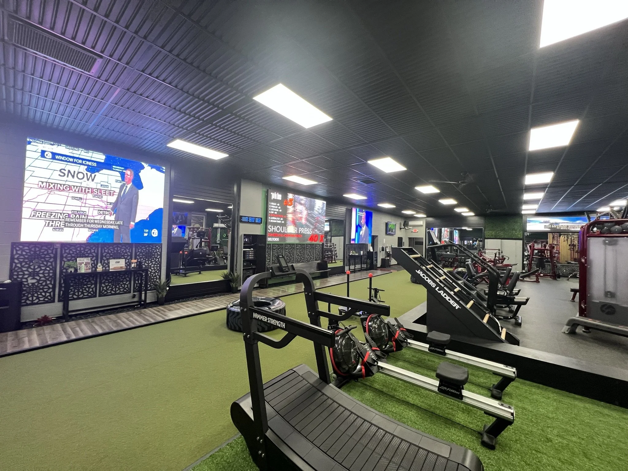 A gym with various workout equipment including benches, weights, and machinery. The ceiling is dark with bright rectangular lights, and there are multiple digital screens displaying information and advertisements. The floor has green turf and pattern