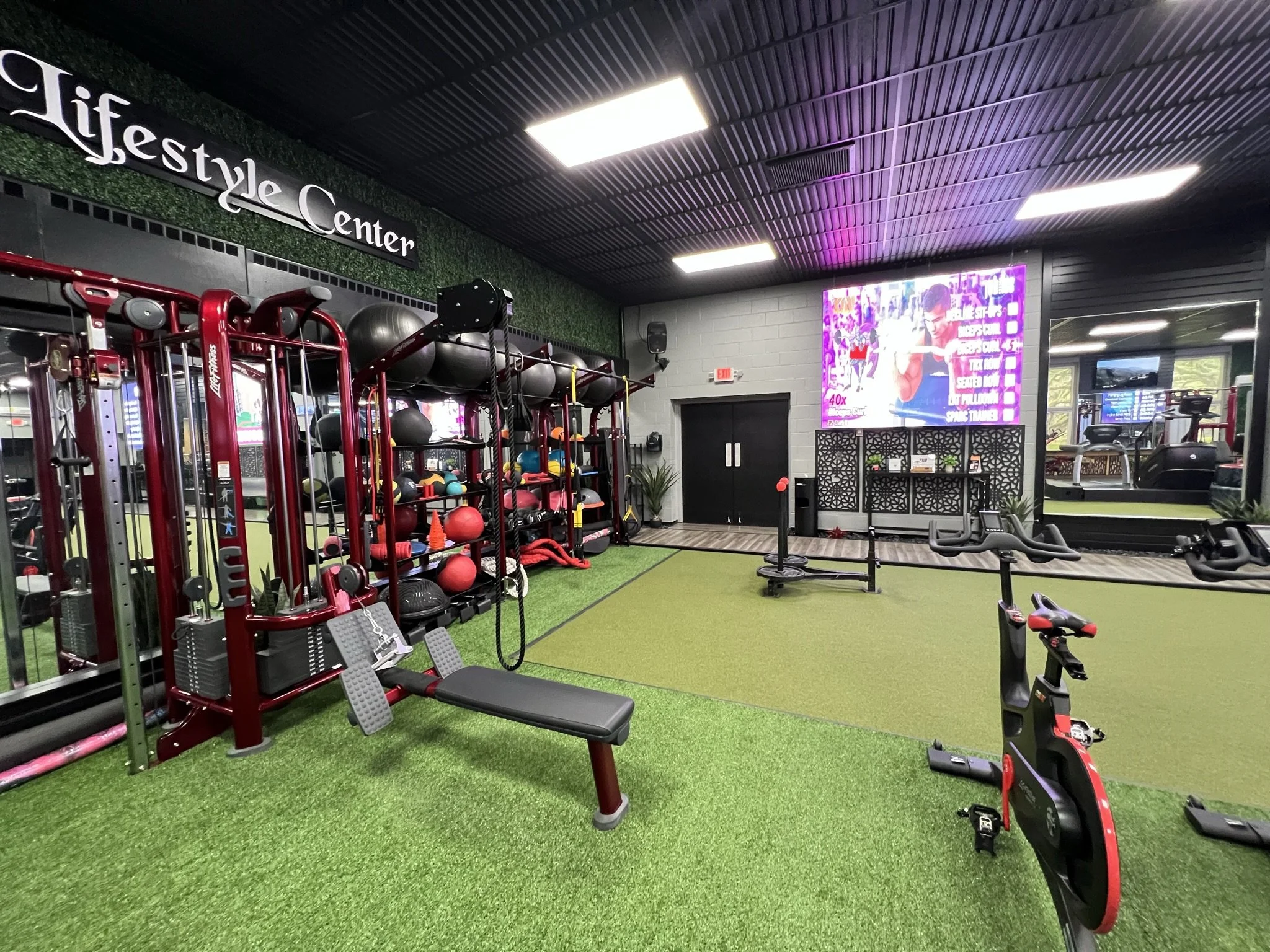 Interior of a gym with fitness equipment, weights, and exercise machines, featuring artificial green turf flooring and a digital screen on the wall.