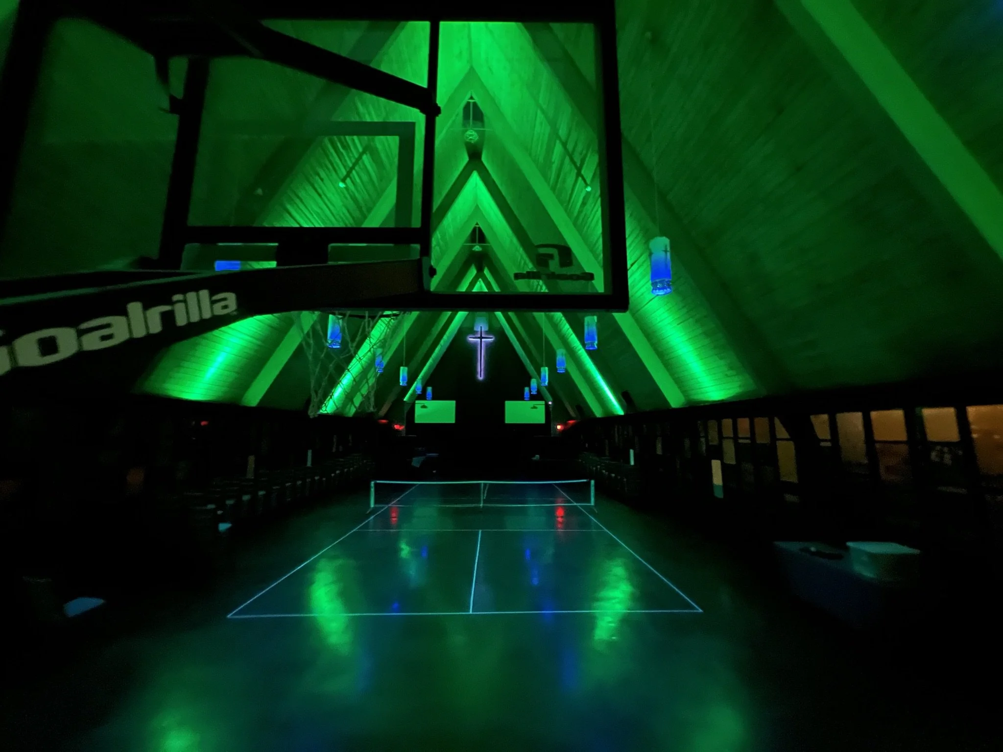 Inside a church with green lighting, an elevated basketball hoop, church cross, and blue hanging lights are visible. The floor is shiny and reflects the lighting.