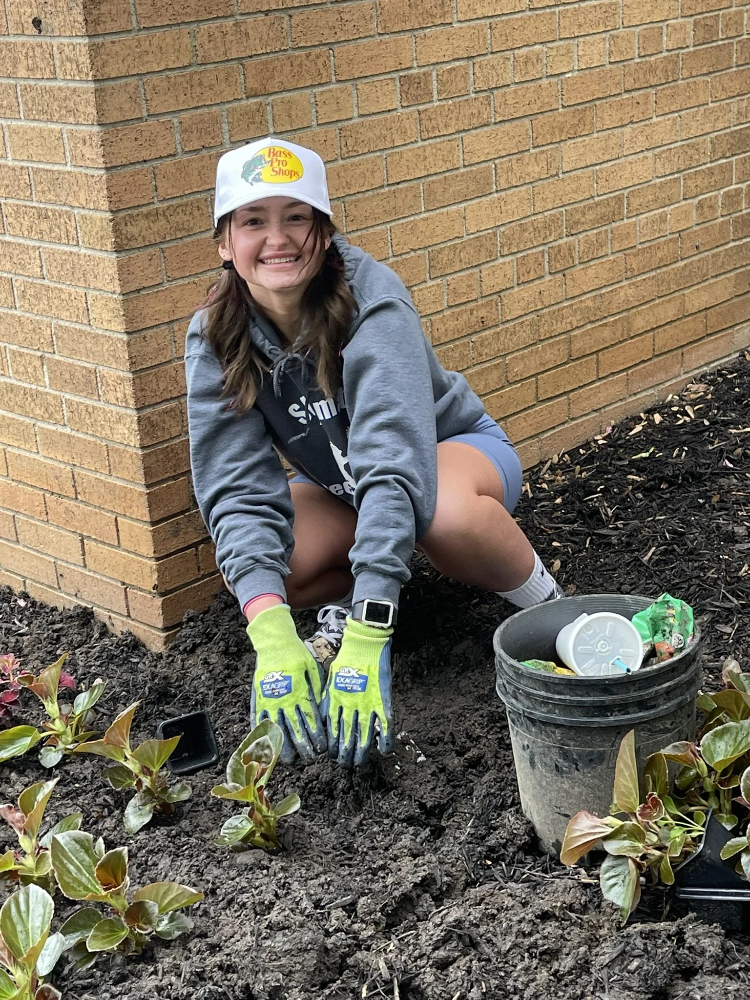 A young woman with long brown hair, wearing a white baseball cap, gray hoodie, and gardening gloves, is smiling while planting flowers near a brick wall. She is crouched down on dark soil with gardening tools and supplies nearby, including a black bu