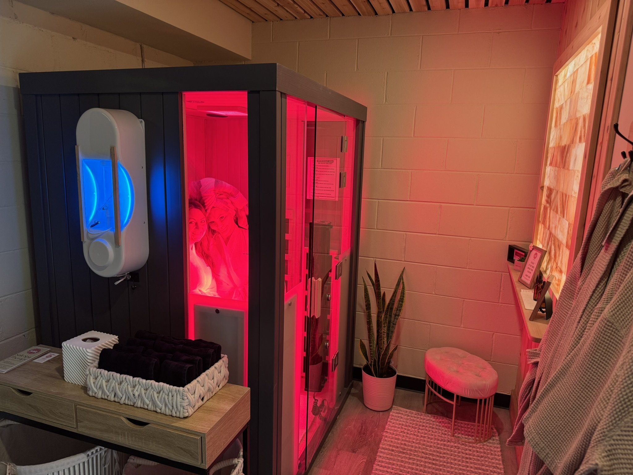 A lighted red infrared sauna inside a room with wood paneling and beige brick walls. Next to it is a bench with towels, a potted plant, and a small pink stool or chair.
