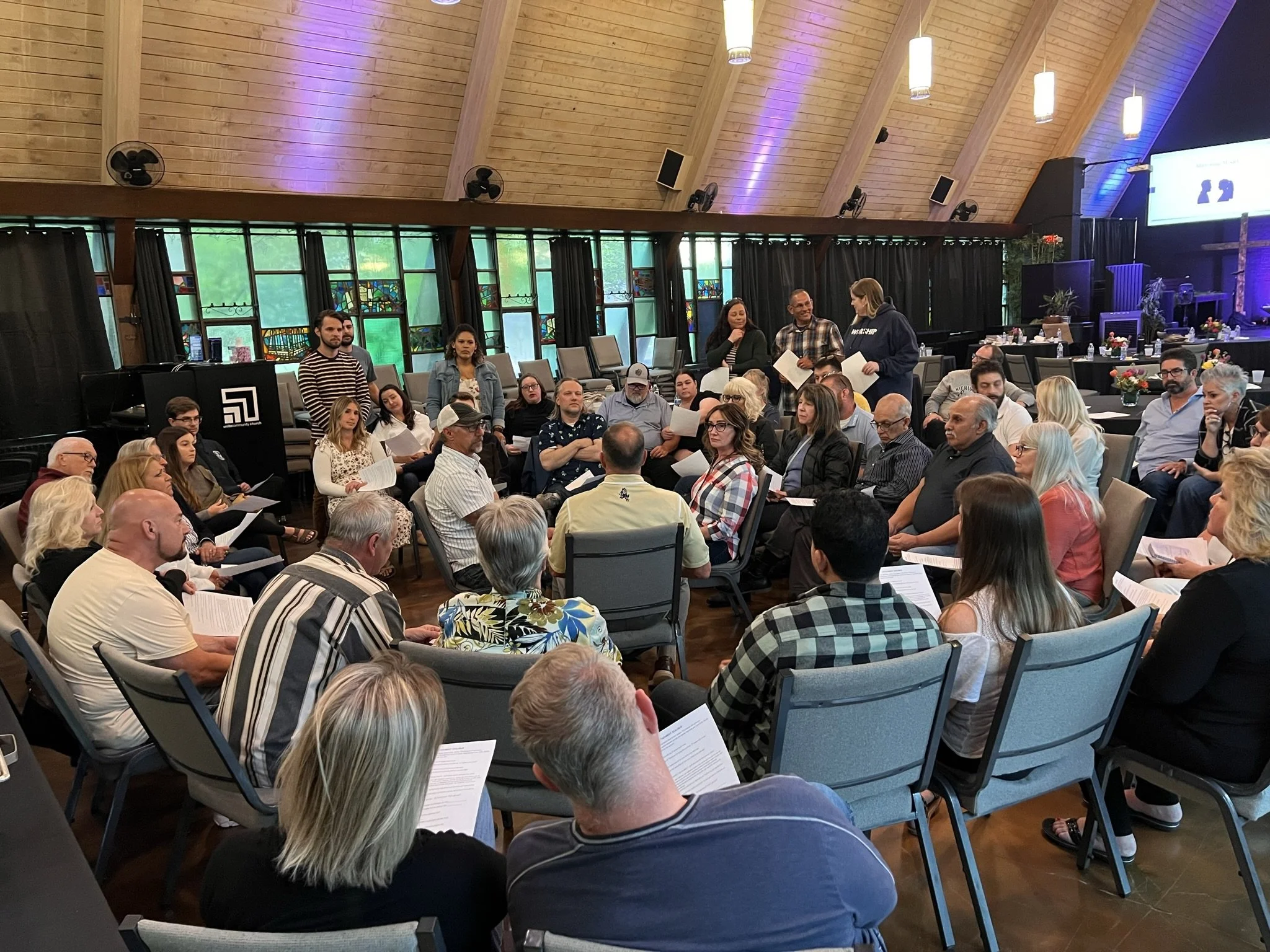 A large group of diverse people seated in a church or conference hall, listening to a presentation or discussion, with some standing and holding papers.