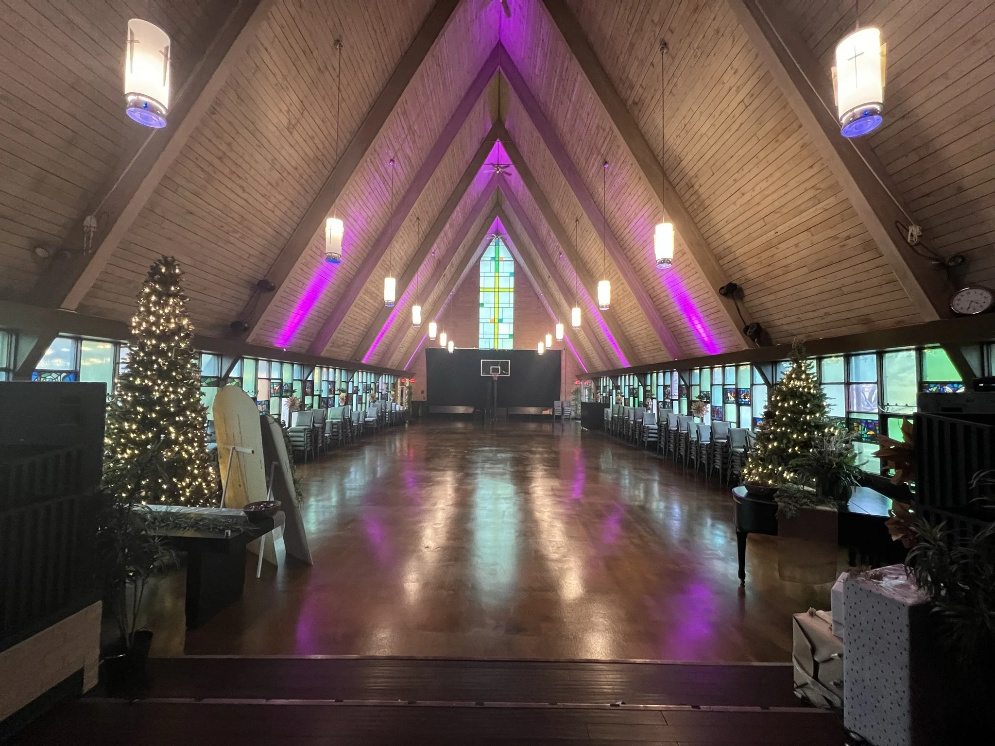 The interior of a church or chapel with high wooden, vaulted ceiling, stained glass windows, Christmas trees with lights, and colorful uplighting on the walls.
