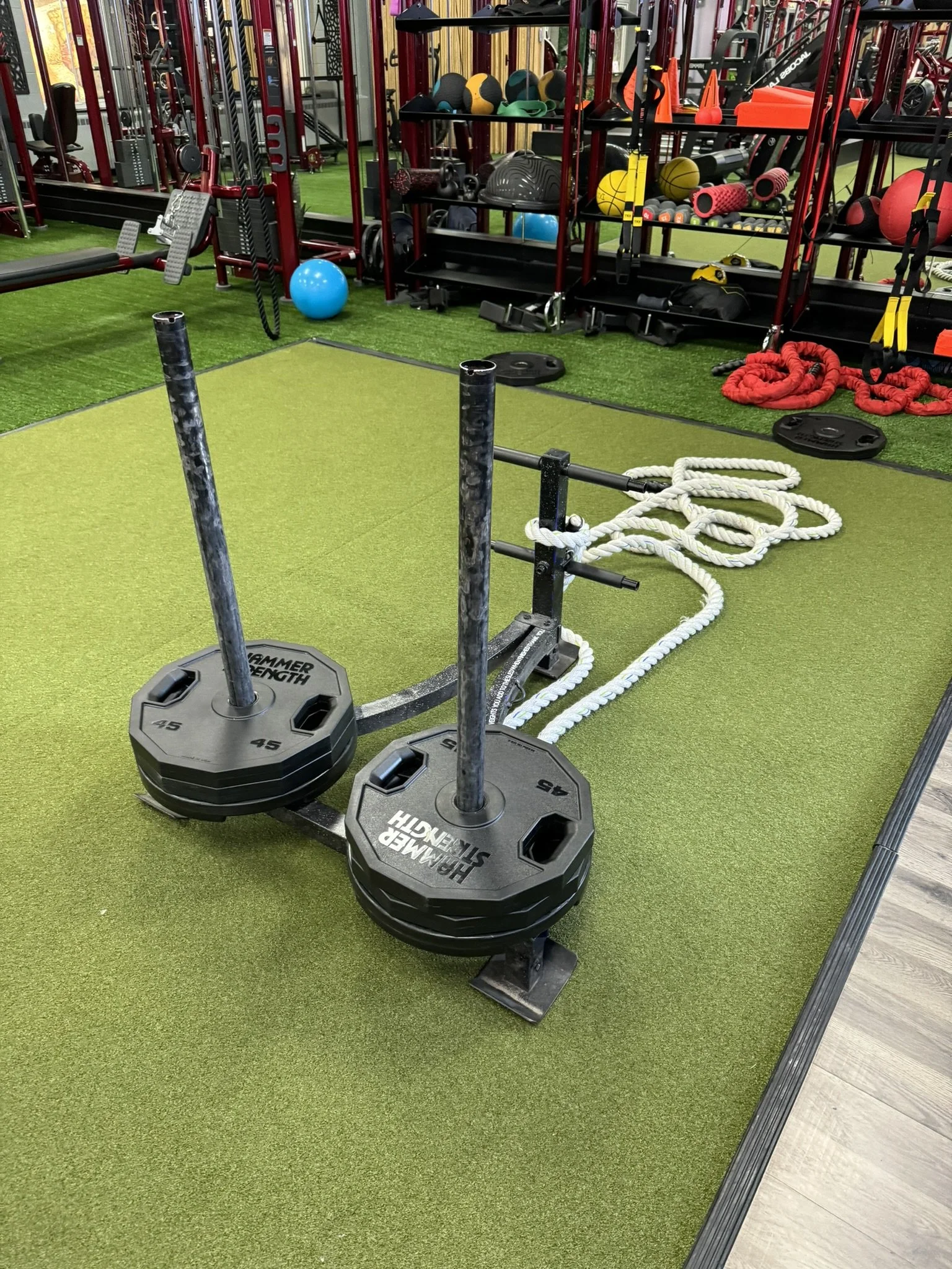 Gym workout area with two barbells on green turf, fitness equipment and accessories in the background.