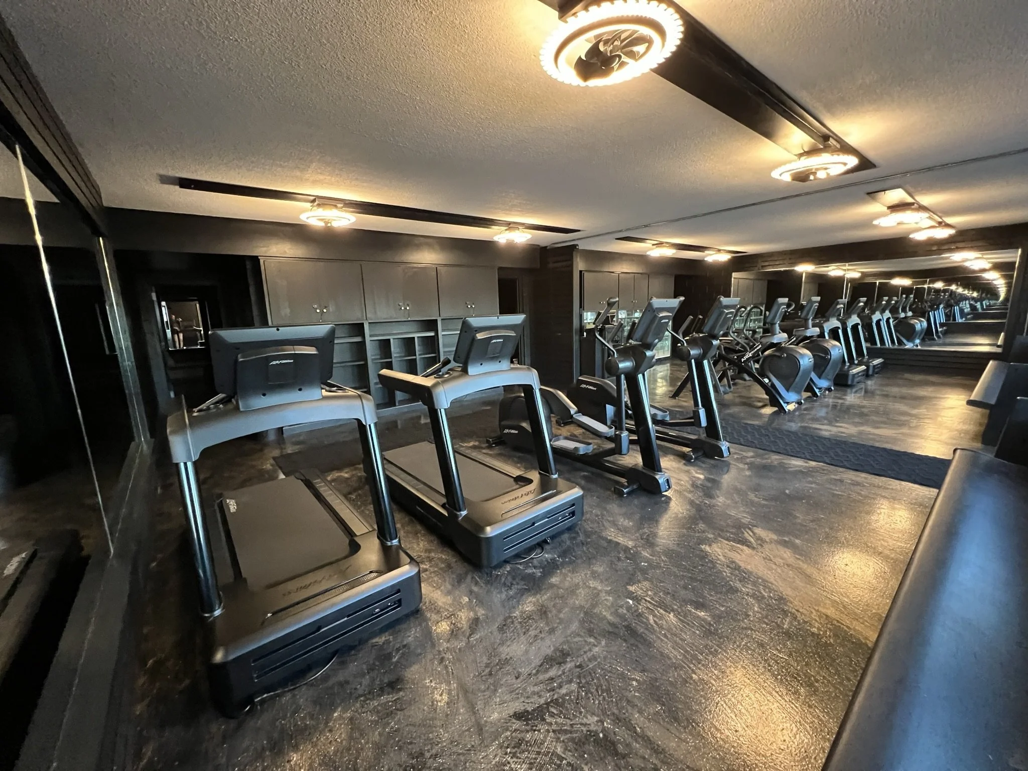 A gym with multiple treadmills lined up in a row, a large mirror on the back wall reflecting the equipment, and ceiling lights illuminating the space.