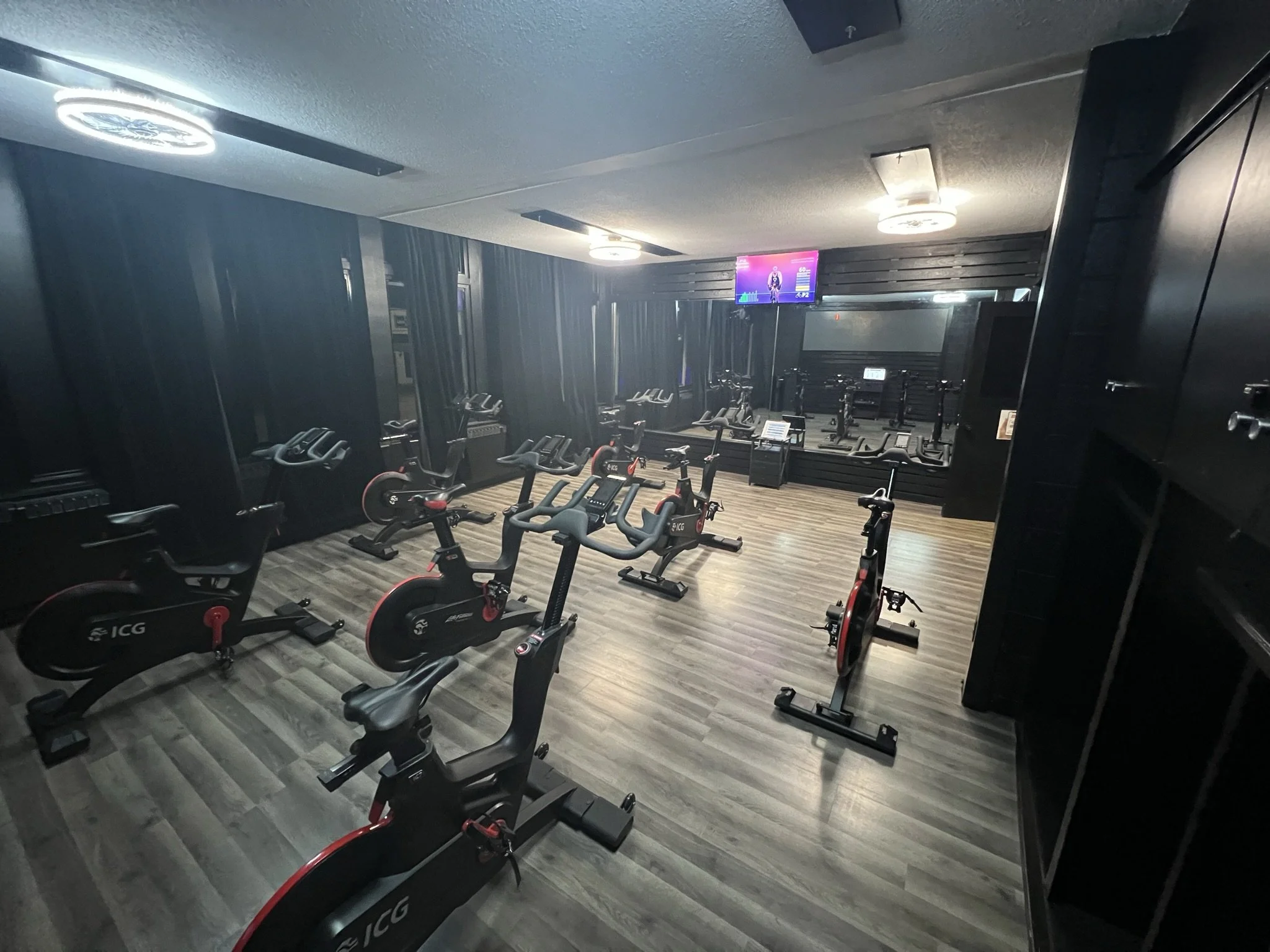 Indoor fitness room with multiple exercise bikes arranged in rows, black walls, wooden floor, and a wall-mounted TV screen.