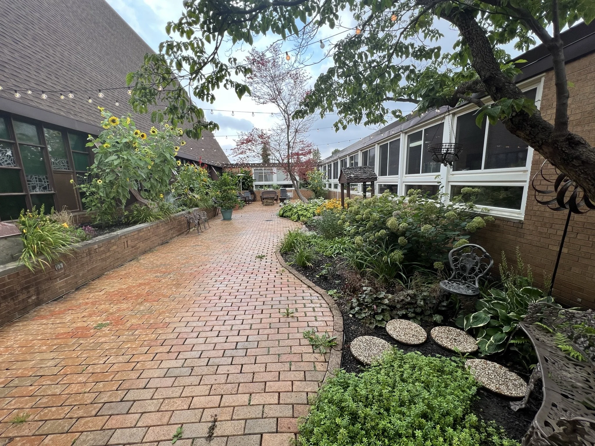A brick patio garden surrounded by plants, flowers, and trees, with string lights hanging overhead, and outdoor seating on a cloudy day.
