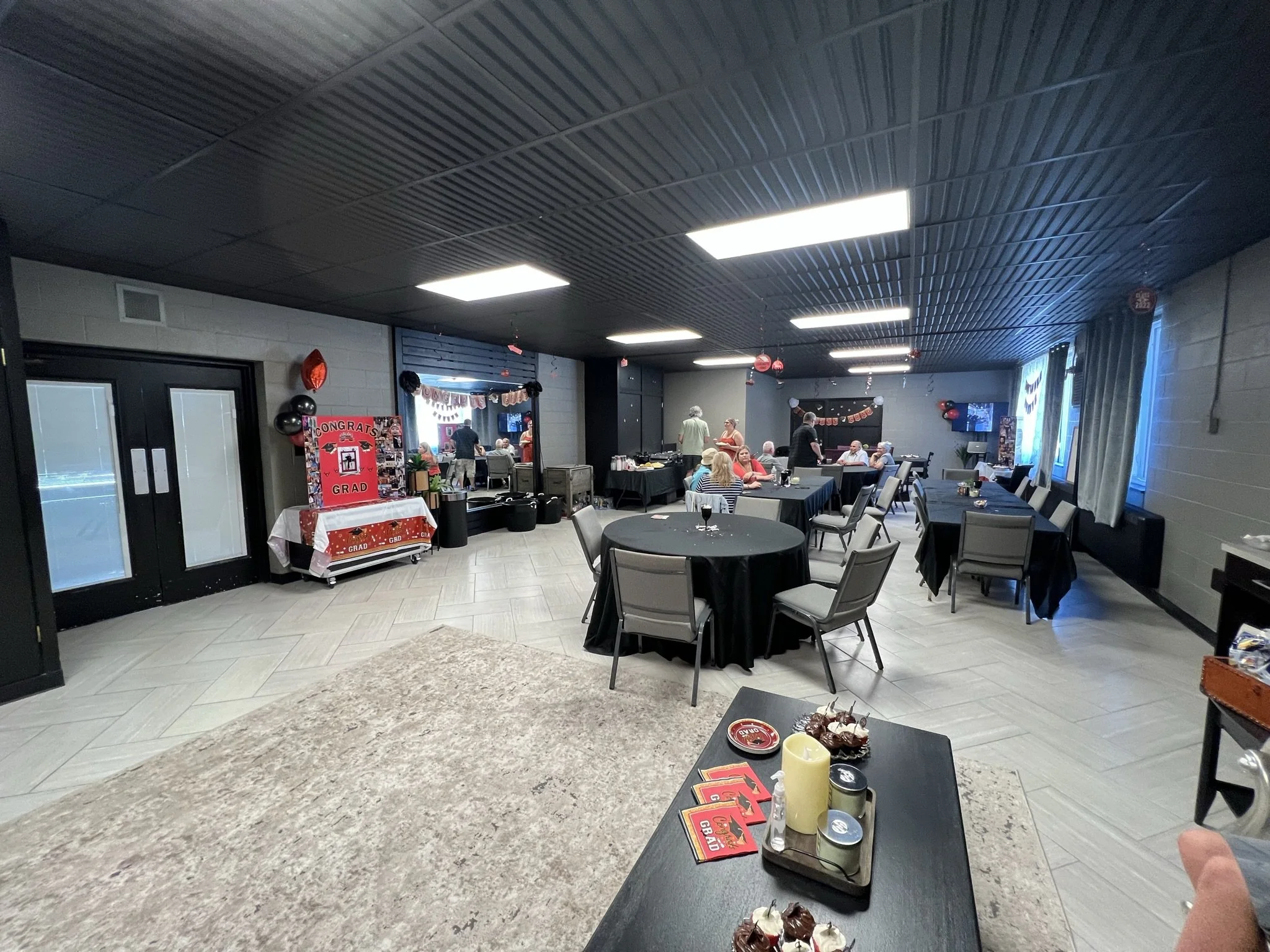 An indoor celebration party with tables, chairs, and decorations, featuring a sign that says 'Congrats Grad' and food on the tables.