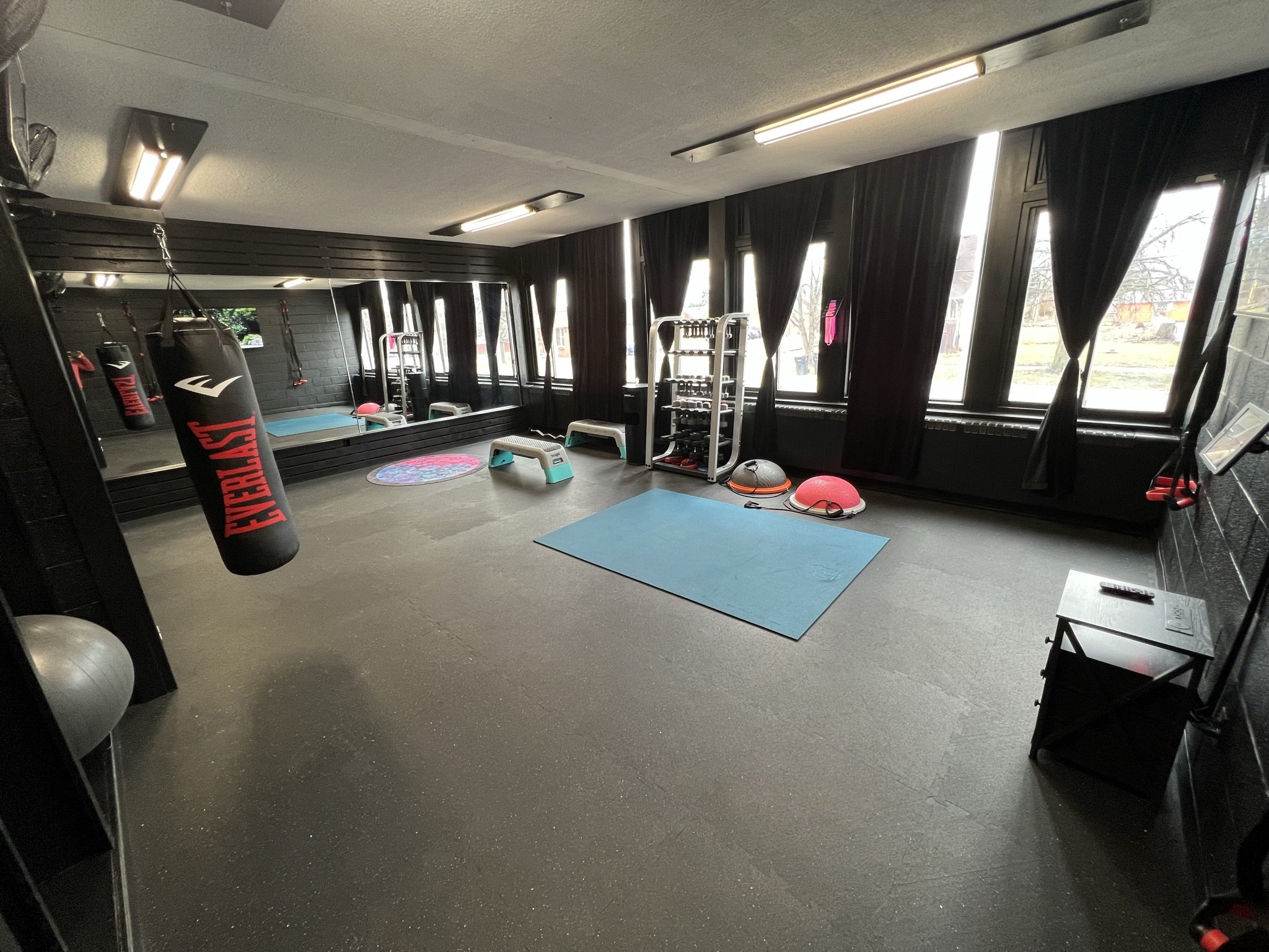 A gym room with cardio and strength training equipment, including punching bags, medicine balls, free weights, step platforms, mats, and large windows with black curtains.