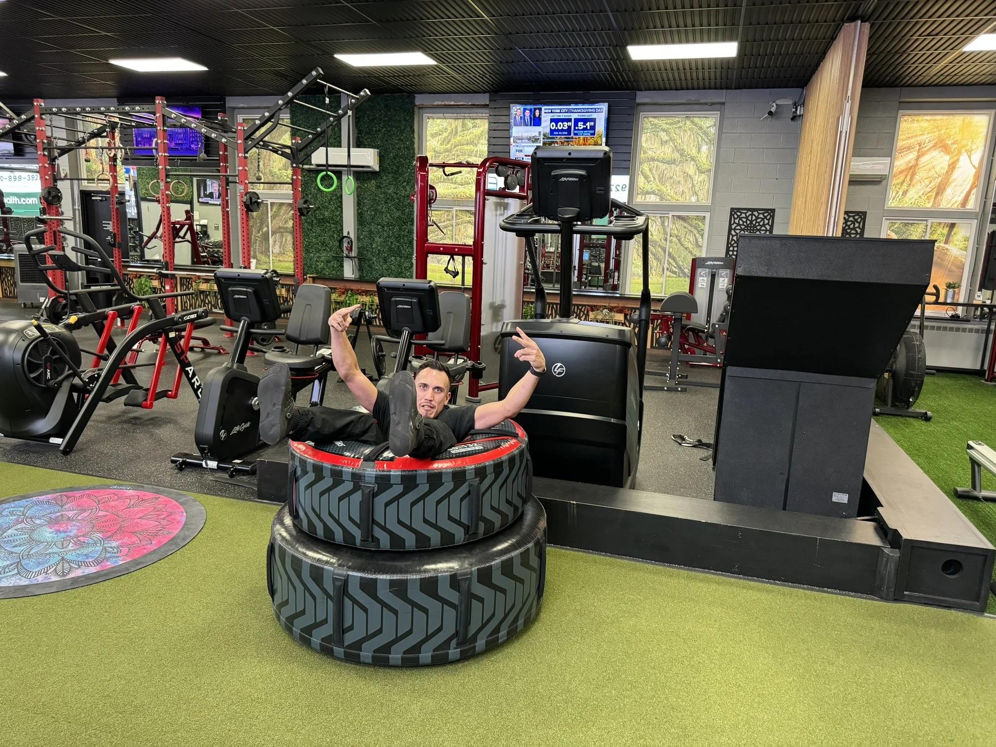 A man lying on a small trampoline in a gym, holding a foam roller and smiling, with gym equipment and large windows showing trees outside in the background.