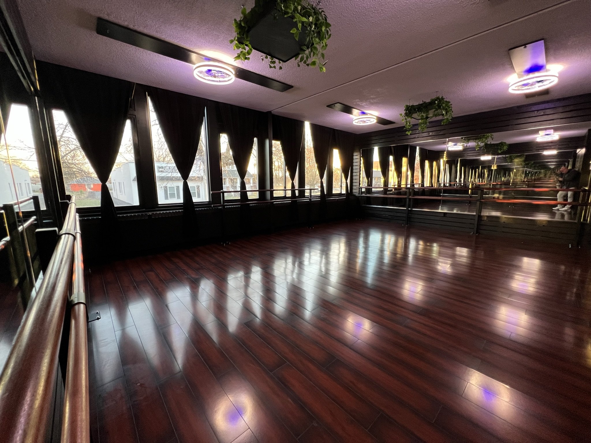 An empty indoor dance studio with large windows, dark curtains, and ceiling lights, featuring wooden floors and ballet barres along the windows.