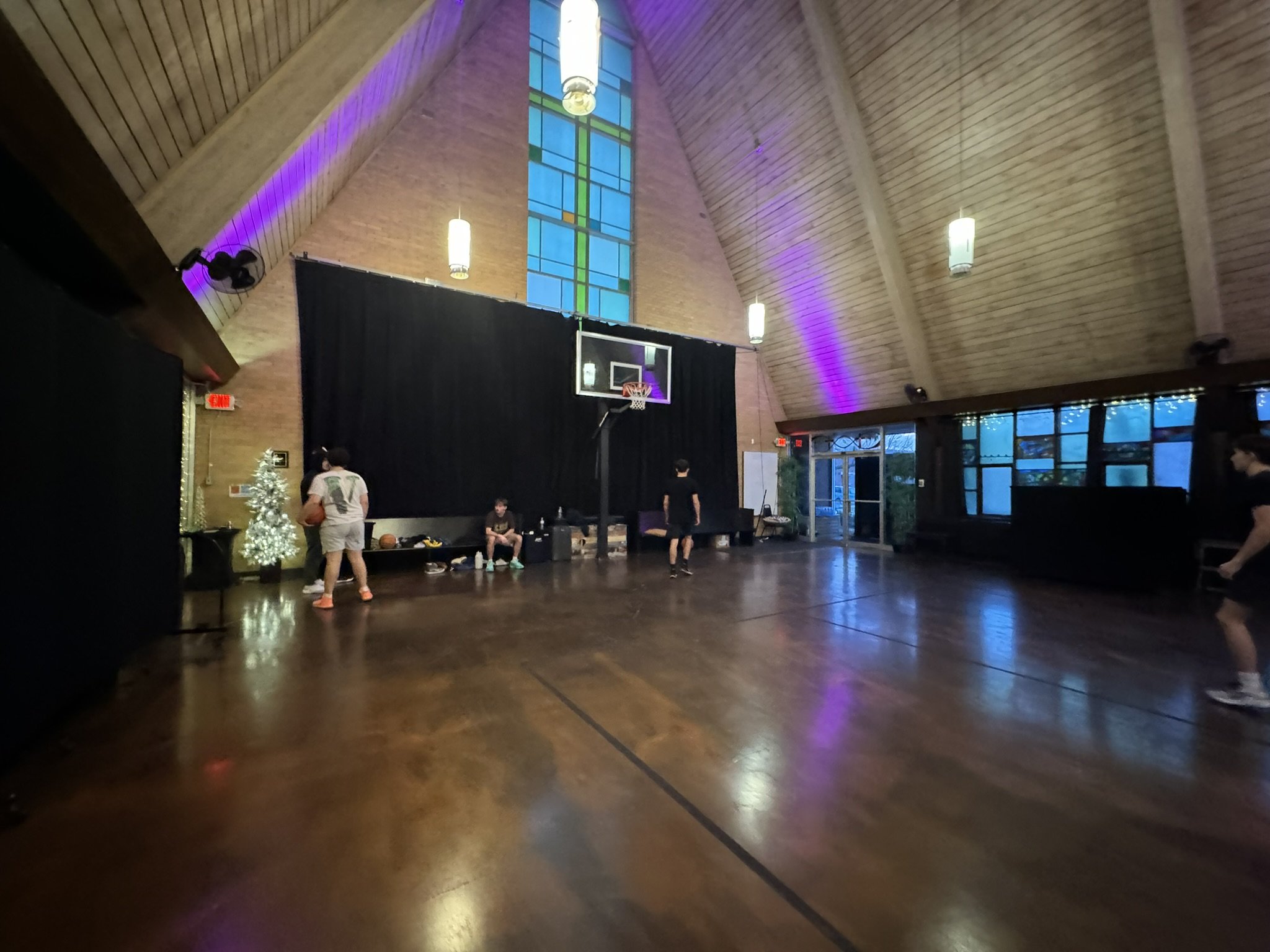 Indoor basketball court with several people, a black curtain, a decorated Christmas tree, and colorful lighting on the walls and ceiling.