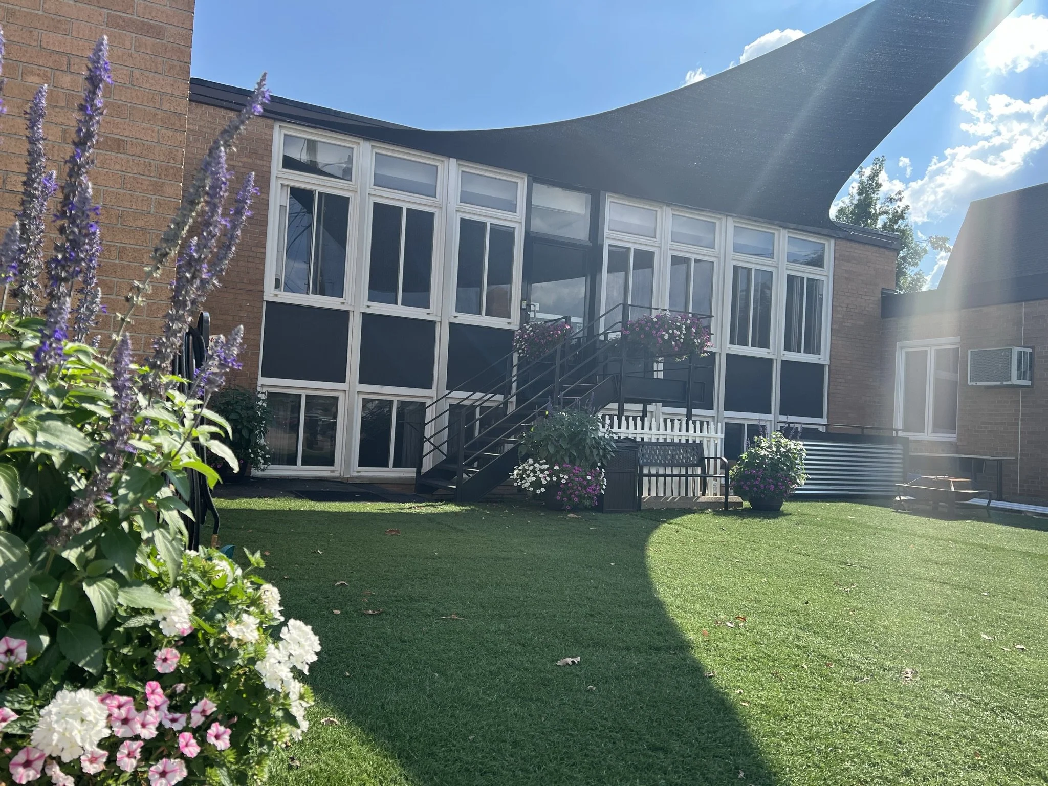 View of a backyard with green artificial grass, garden flowers in the foreground, and a brick house with a screened porch and large windows in the background, bright sunny day.