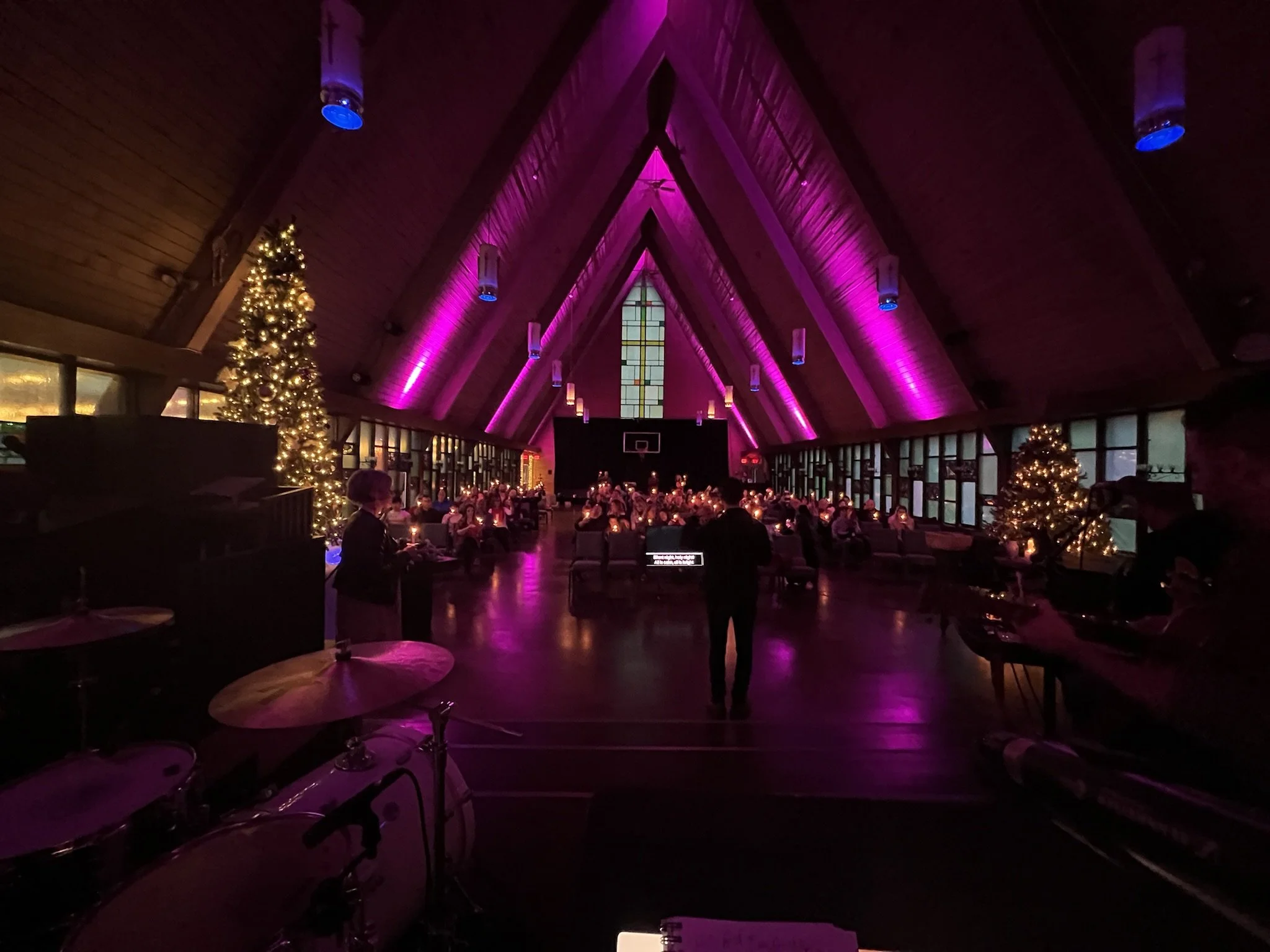 A church hall decorated for Christmas with Christmas trees and purple lighting, filled with seated people and hanging cylindrical lights.