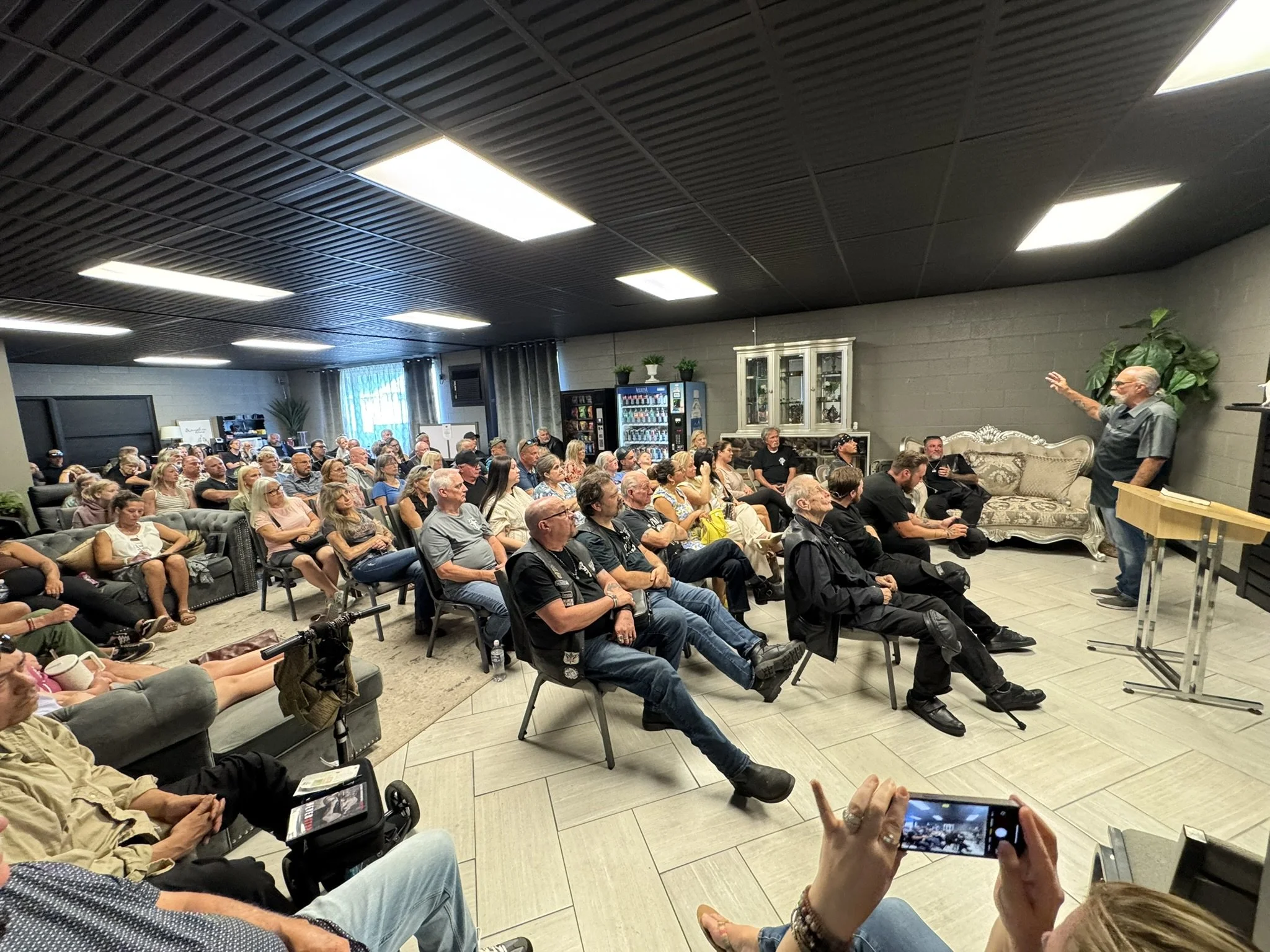 A large group of people seated in a room, listening to a speaker standing next to a sofa against the wall, with some individuals taking photos or recording the event.