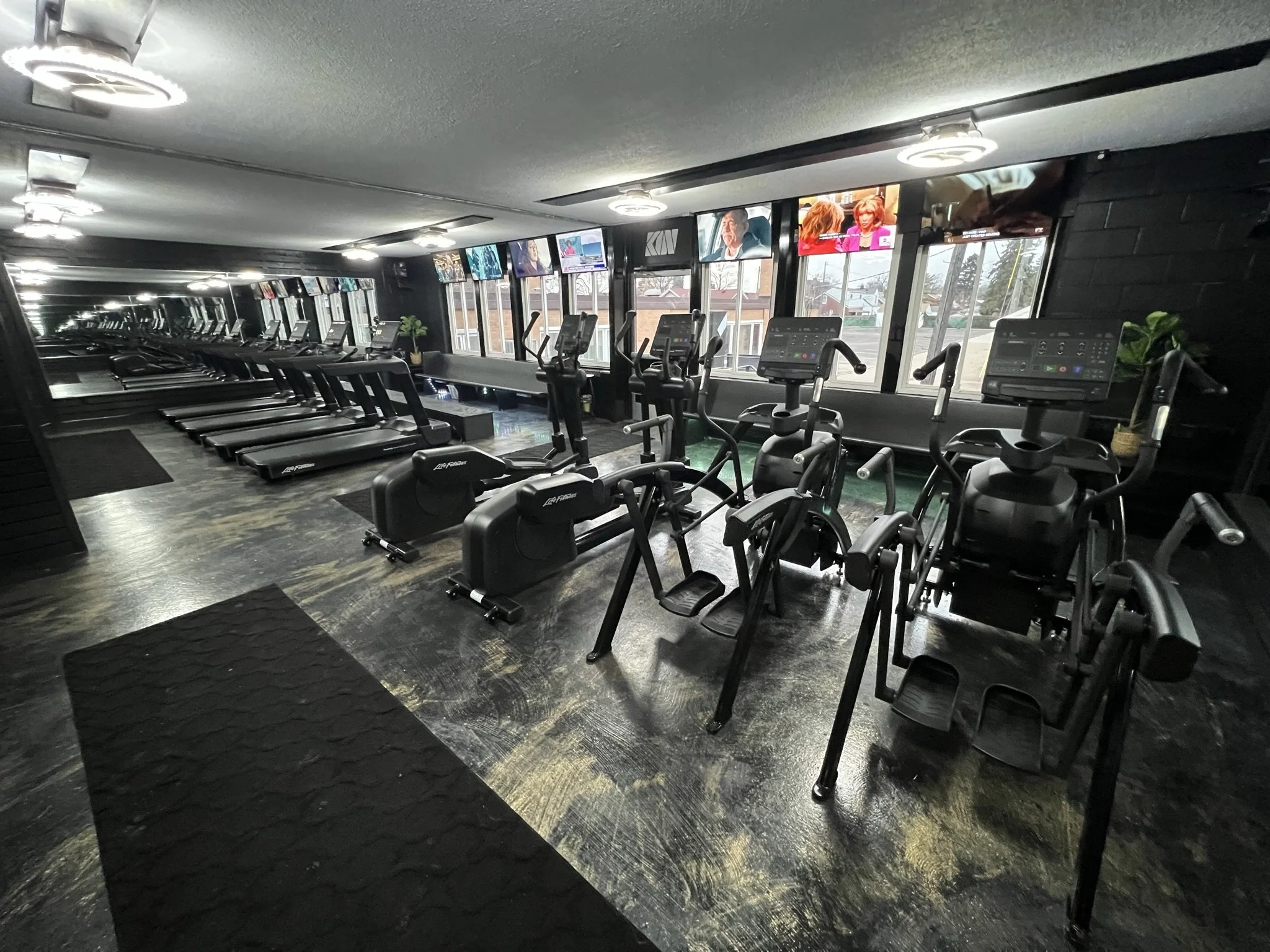 A modern gym with treadmills, ellipticals, and exercise bikes facing large windows. Multiple TV screens are mounted above the machines. The floor is dark with rubber mats, and interior lighting is bright.
