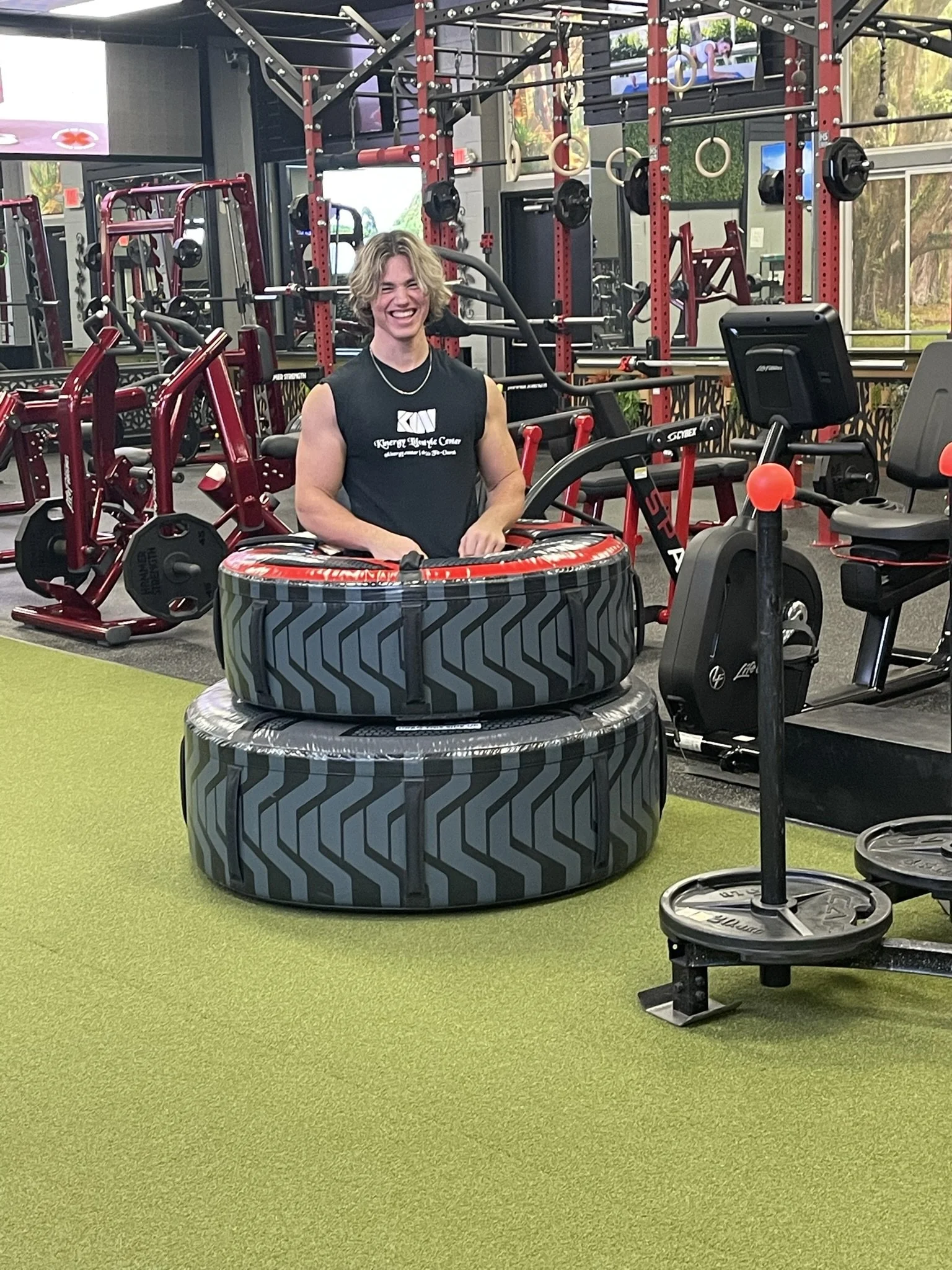 A young man with blonde hair smiling in a gym surrounded by fitness equipment, including large tires, weight machines, and a rowing machine.
