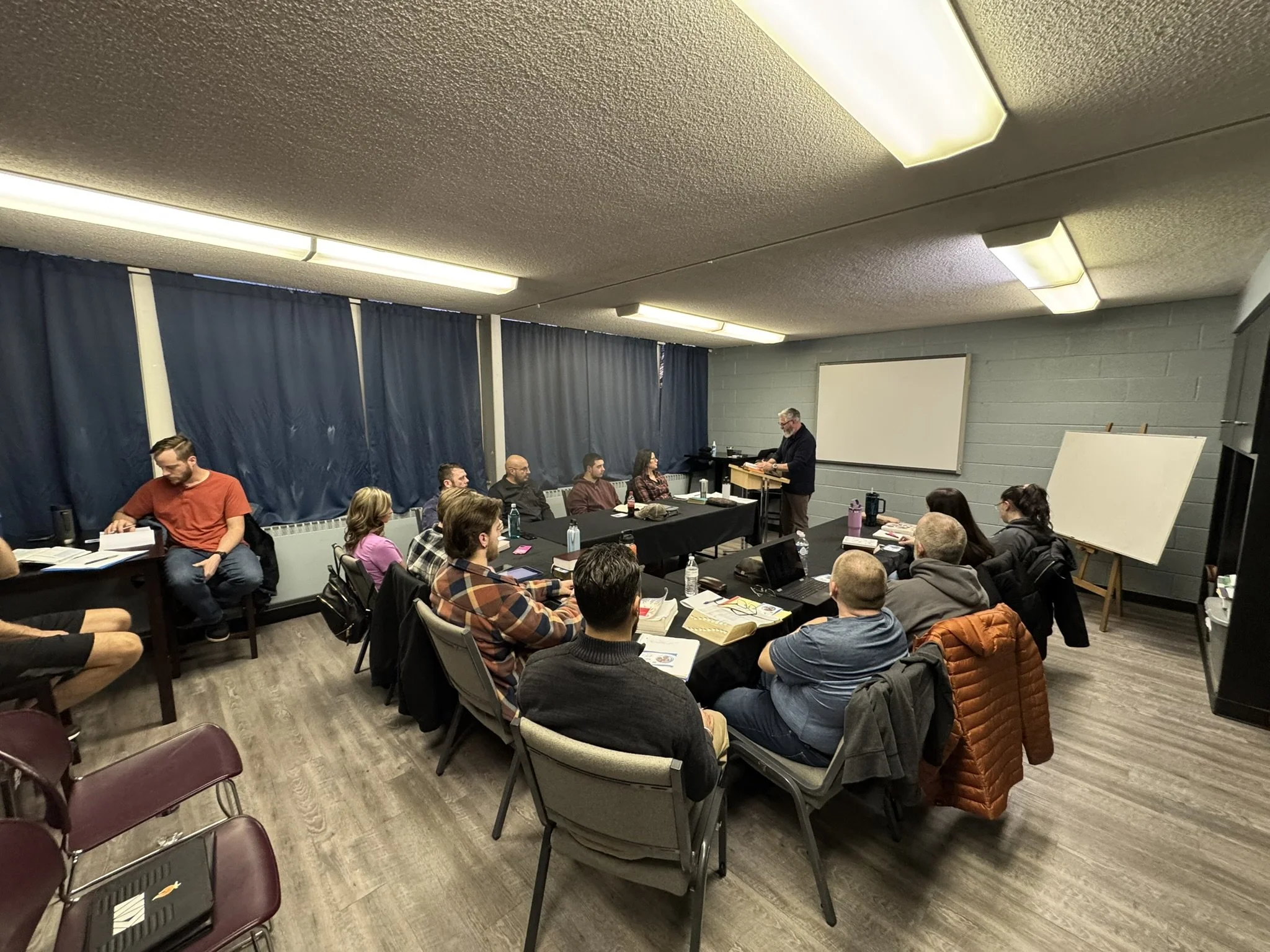 A group of people attending a seminar or workshop in a conference room, seated around tables with notebooks, water bottles, and laptops, listening to a speaker standing at the front of the room.