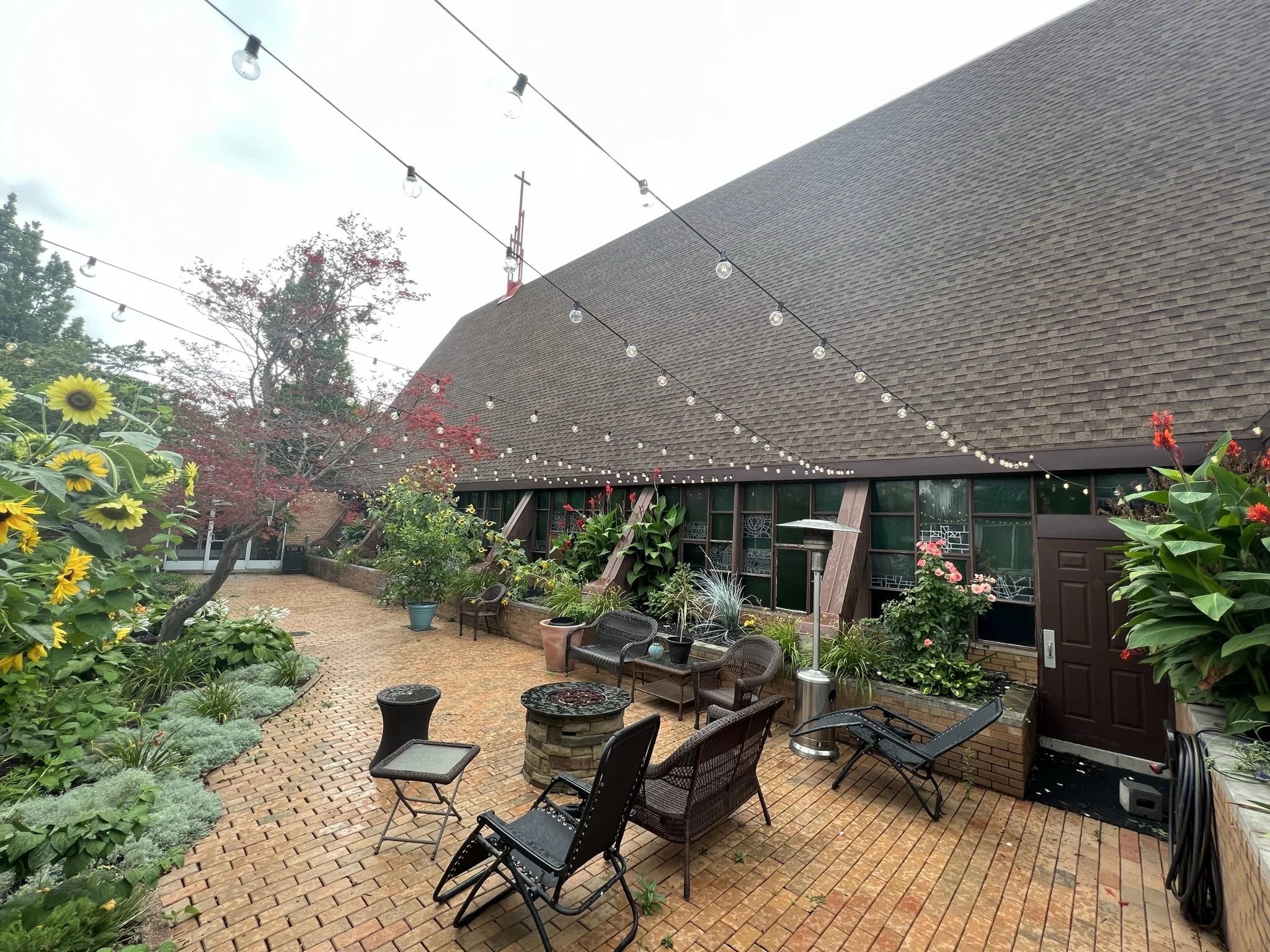 A backyard patio with brick flooring, surrounded by colorful flowers and plants. String lights hang overhead, and patio furniture is arranged around a fire pit. There is a building with large windows and a roof with a church steeple visible above.