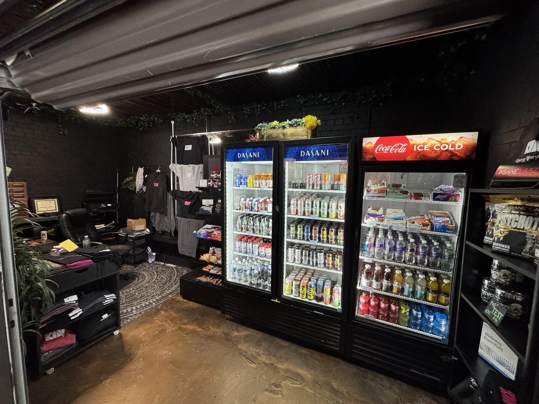 An indoor convenience store with black walls and ceiling, featuring refrigerators stocked with beverages, racks of clothing, and various snacks on shelves, with a patterned rug on the wooden floor.