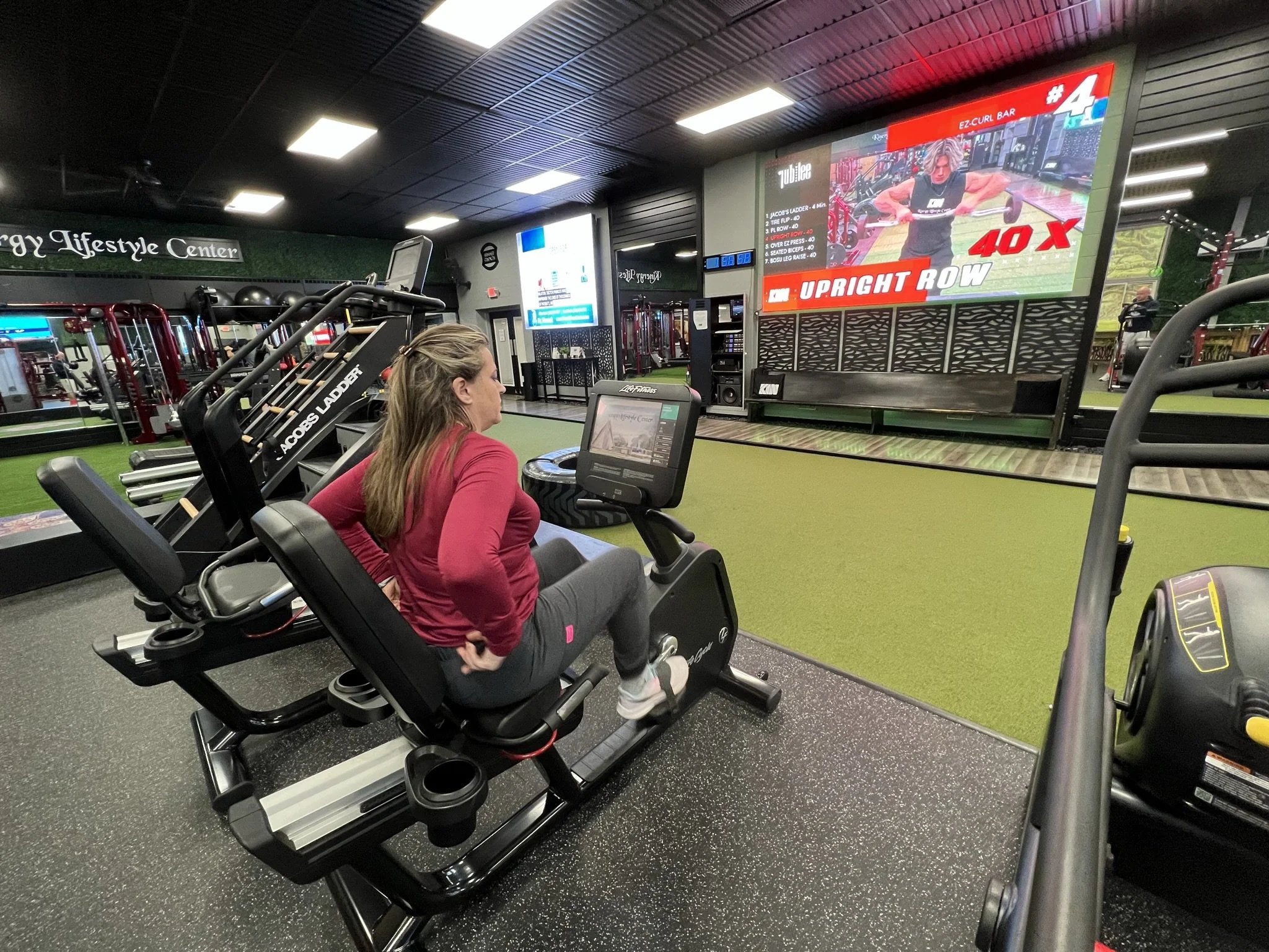 A woman in a red jacket exercising on a stationary bike inside a gym. In the background, a large screen displays an exercise video and a list of workout instructions. The gym has black ceiling panels and various workout equipment.