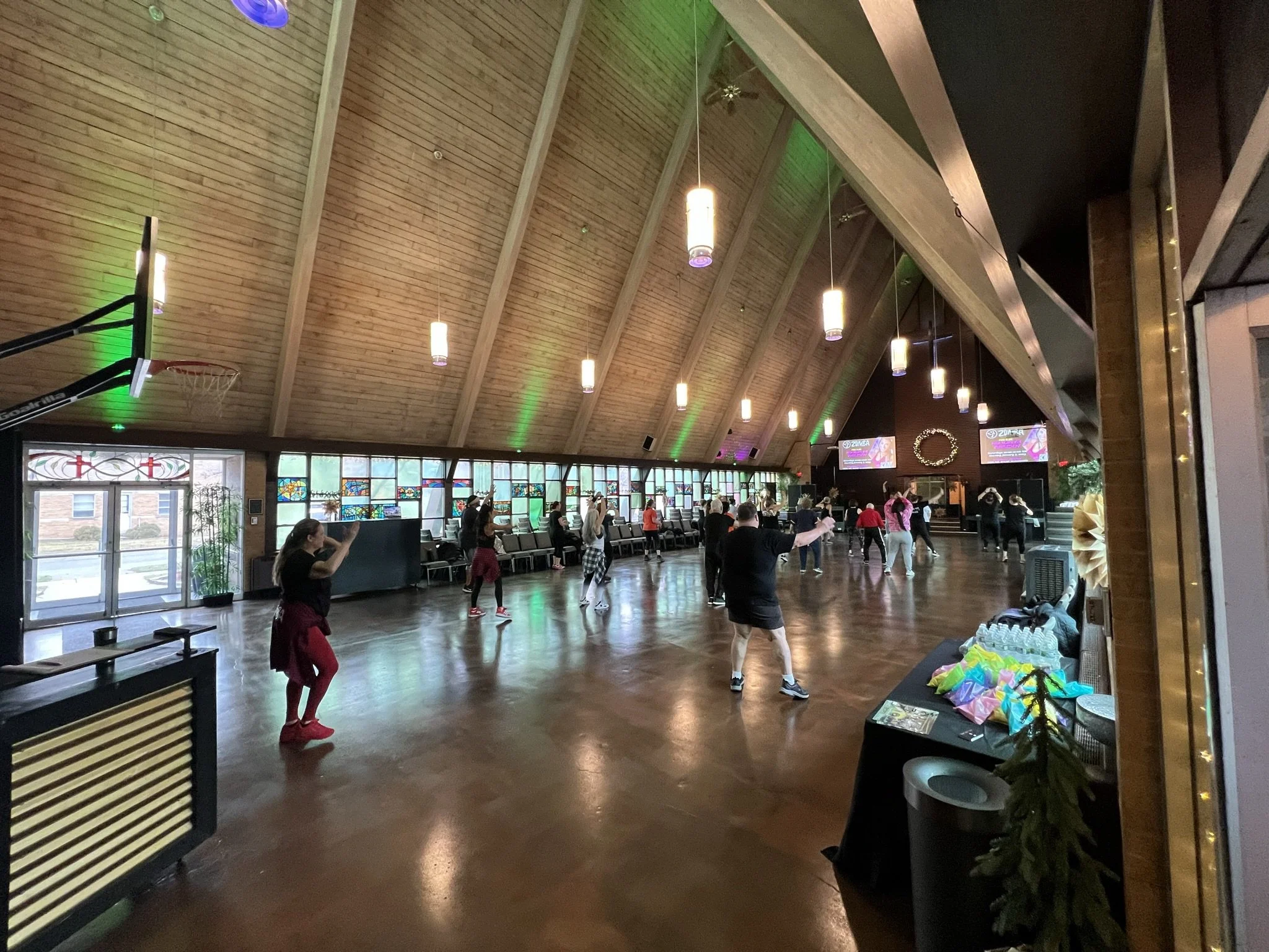 People participating in a dance or exercise class inside a spacious room with a high vaulted wooden ceiling, large windows with stained glass, and hanging pendant lights.