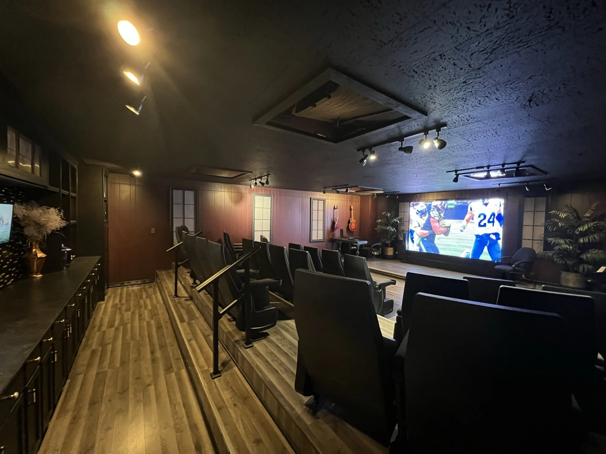 Living room with a large flat-screen TV showing people in football uniforms, several black chairs, wooden floor, wall-mounted guitars, plants, and ceiling lights.