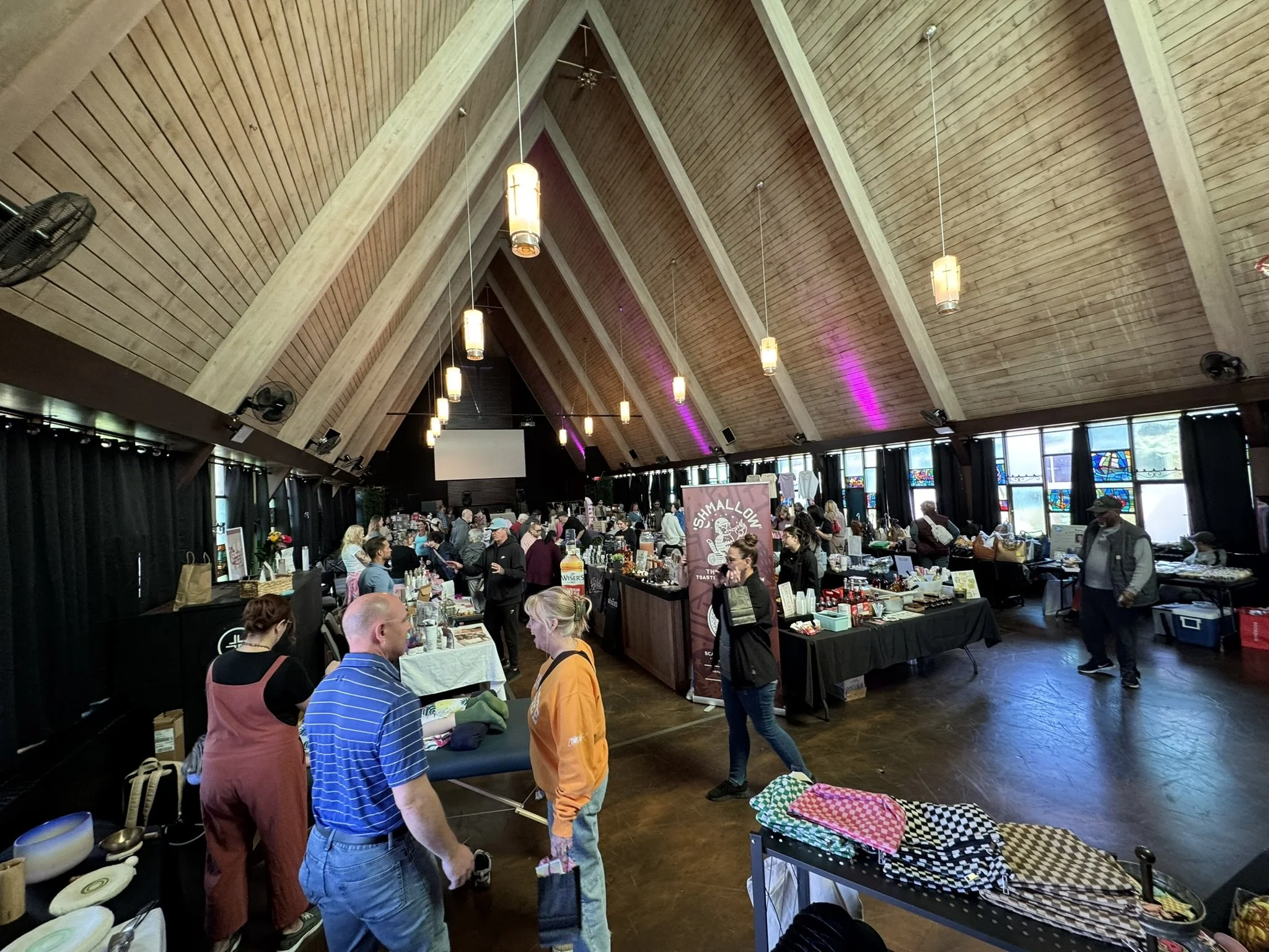 Indoor event with people browsing vendor tables and displays in a large, high ceiling; wooden beams and hanging lights.