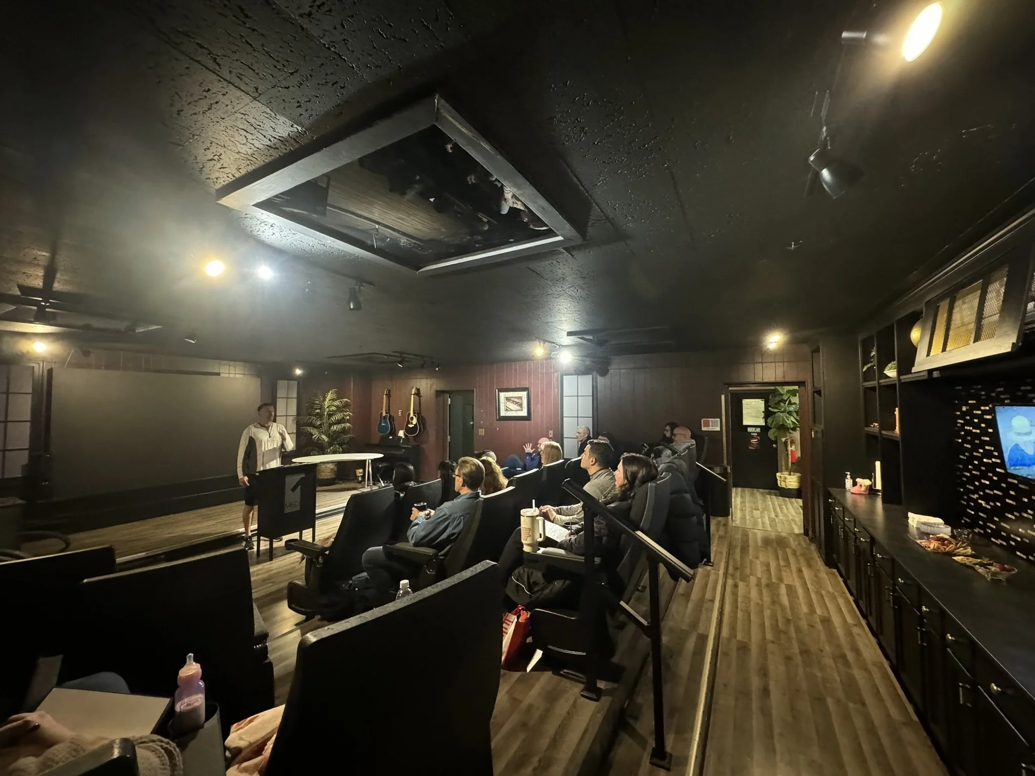 Indoor event with an audience seated in black chairs, a performer with a microphone on stage, and guitars hanging on the wall. The ceiling has a square opening with people seen through it, and dim lighting creates a cozy atmosphere.