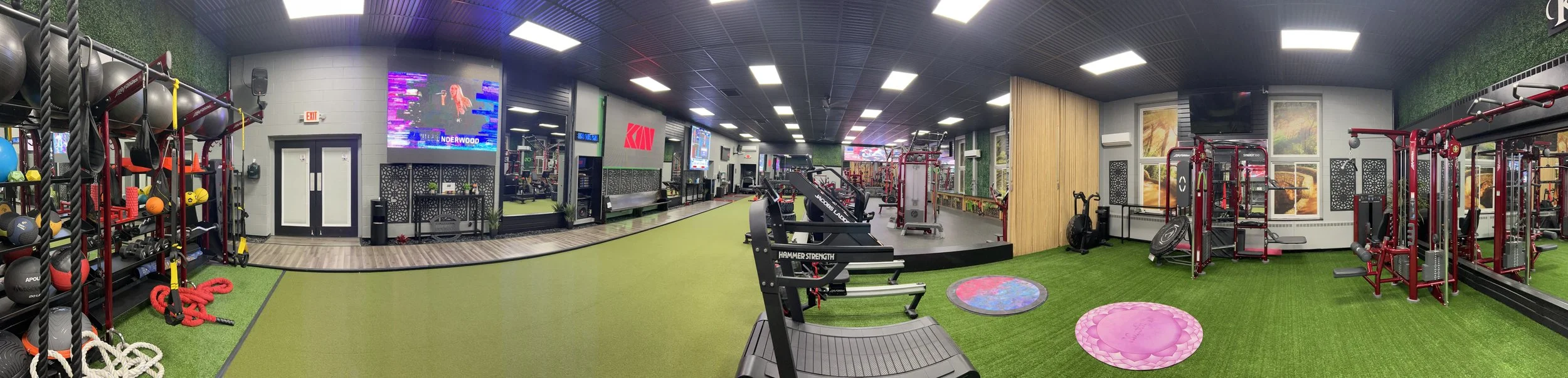 Interior view of a gym featuring weightlifting equipment, cardio machines, and a media wall.