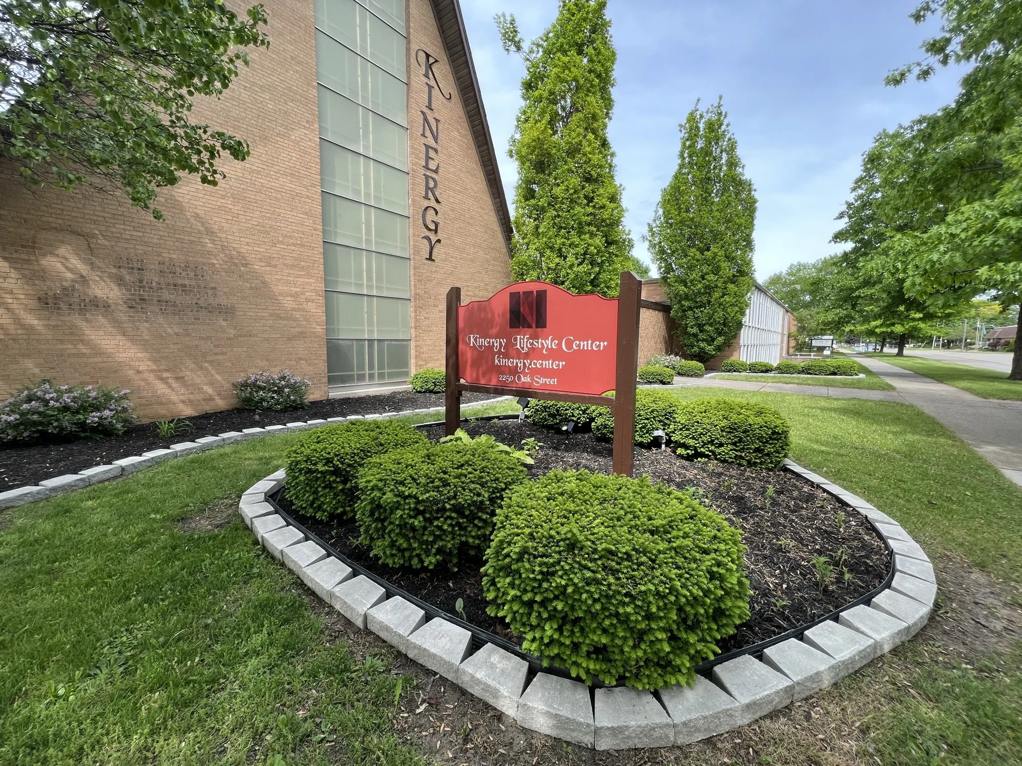 The image shows the exterior of the Kinergy Lifestyle Center building, featuring a large glass window and a brick wall. In front, there is a landscaped flower bed with neatly trimmed bushes, flower plants, and a border made of gray bricks. A red sign with white and black text indicates the center's name and address, situated on a well-maintained grassy area. There are trees and a sidewalk along the street in the background.