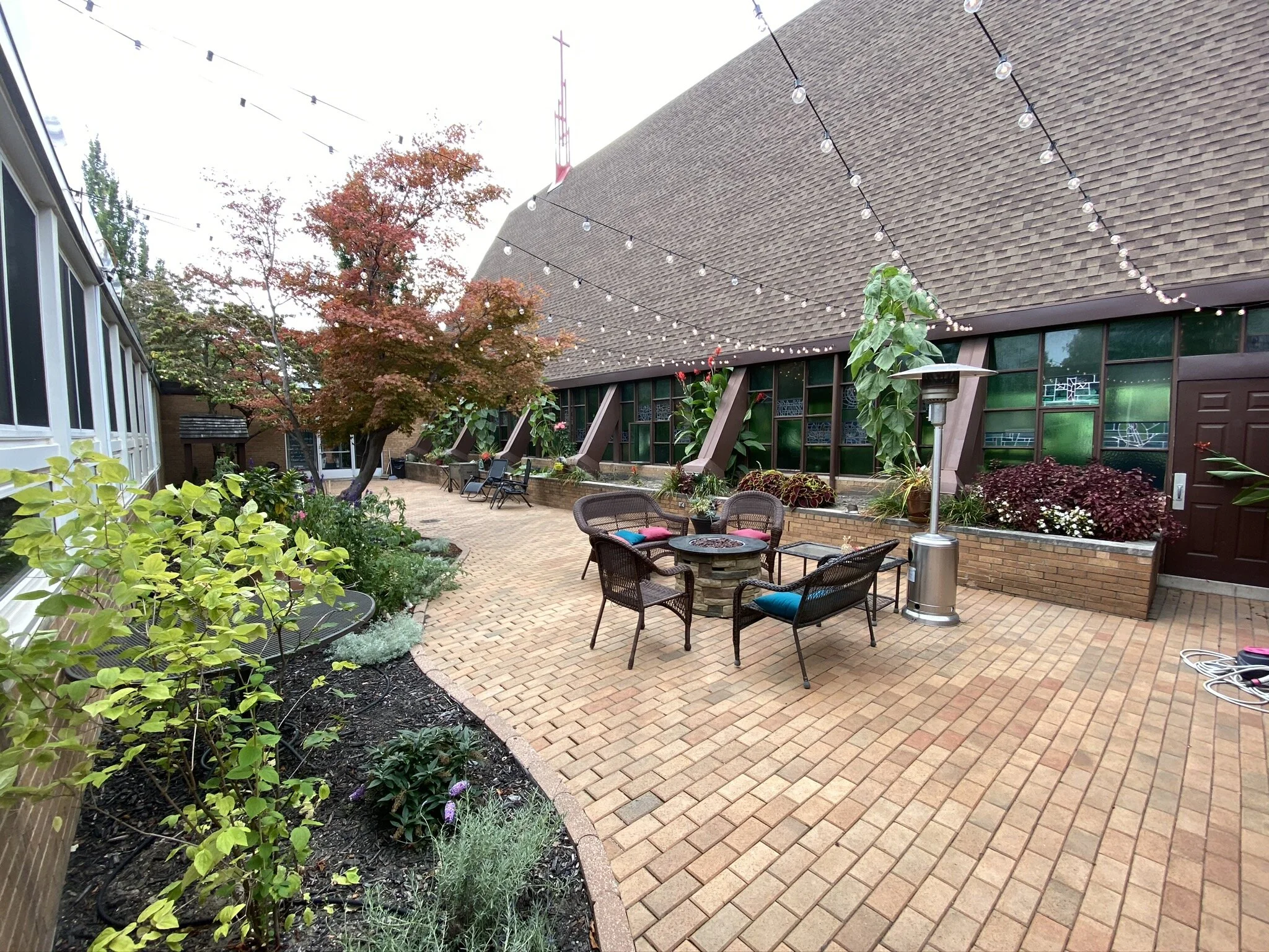 Outdoor patio area with brick paving, surrounded by green plants, trees with autumn leaves, and a building with large windows. String lights are hung overhead, and patio chairs and tables are arranged nearby.