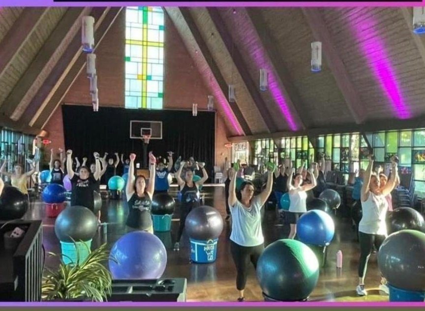 Group fitness class taking place in a church-like gym with high wooden ceilings, stained glass windows, and purple lighting. Participants are doing aerobics with exercise balls in front of a stage.