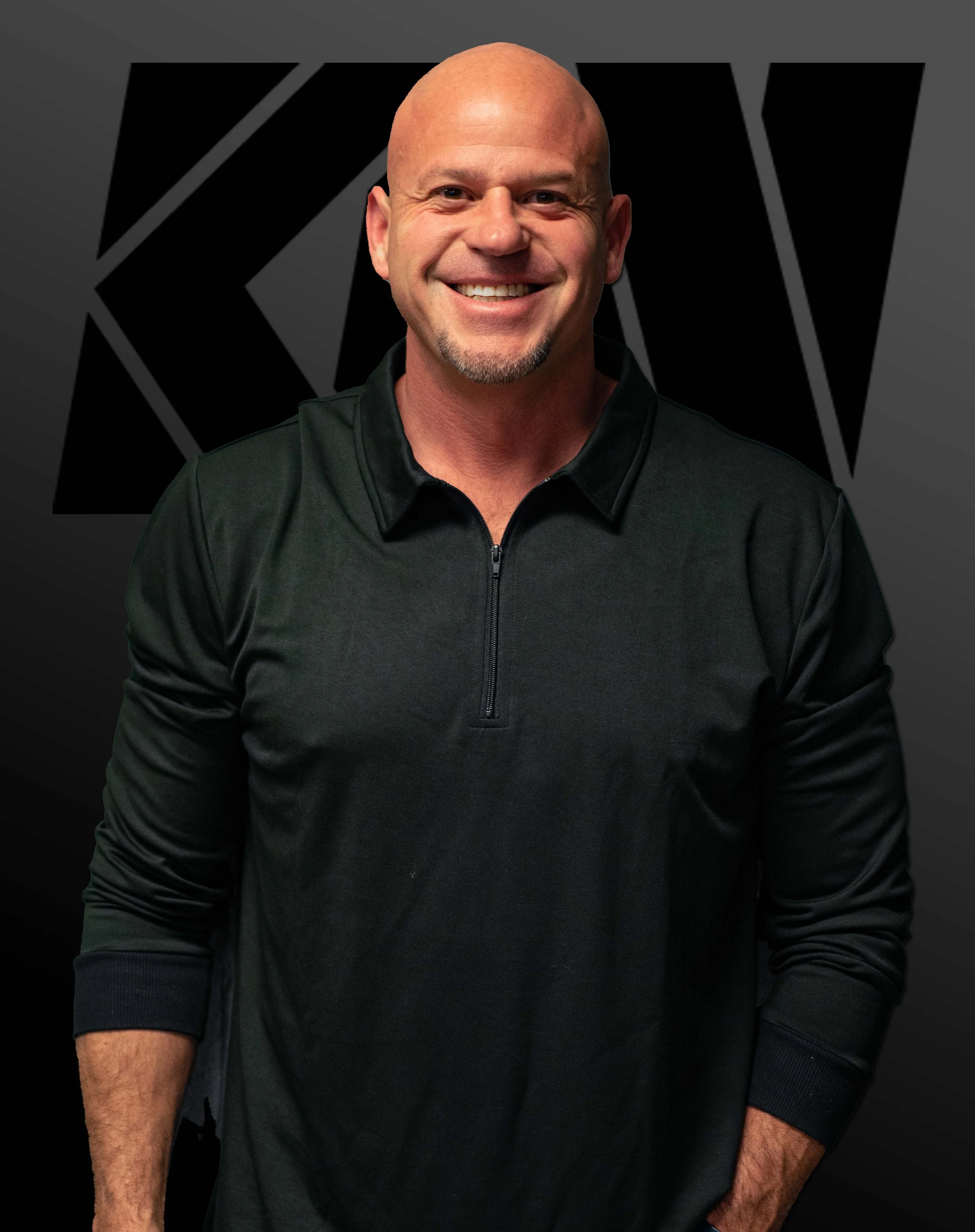 A smiling bald man with a goatee, wearing a black zip-up sports jacket, standing in front of a dark background with a black and silver geometric design.
