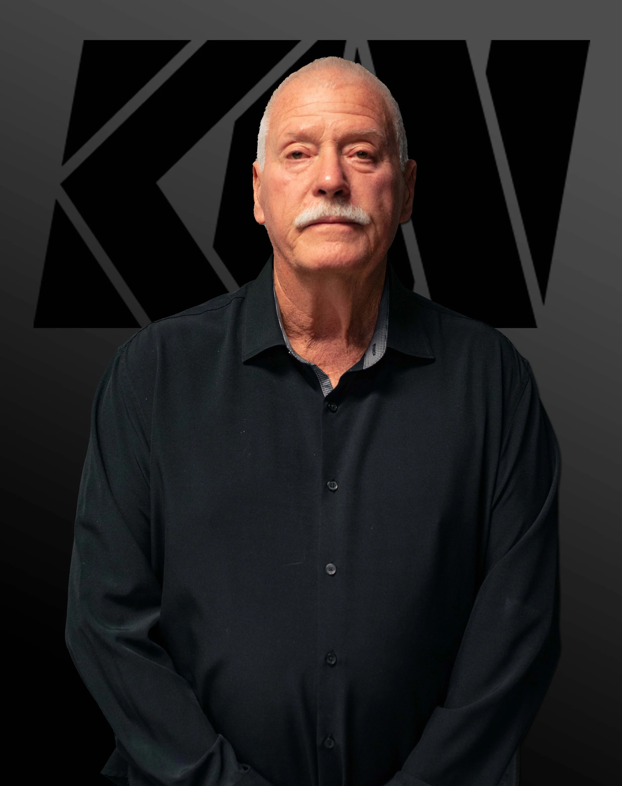 A middle-aged man with a mustache wearing a black dress shirt, sitting with arms crossed, in front of a dark background with an abstract geometric design.