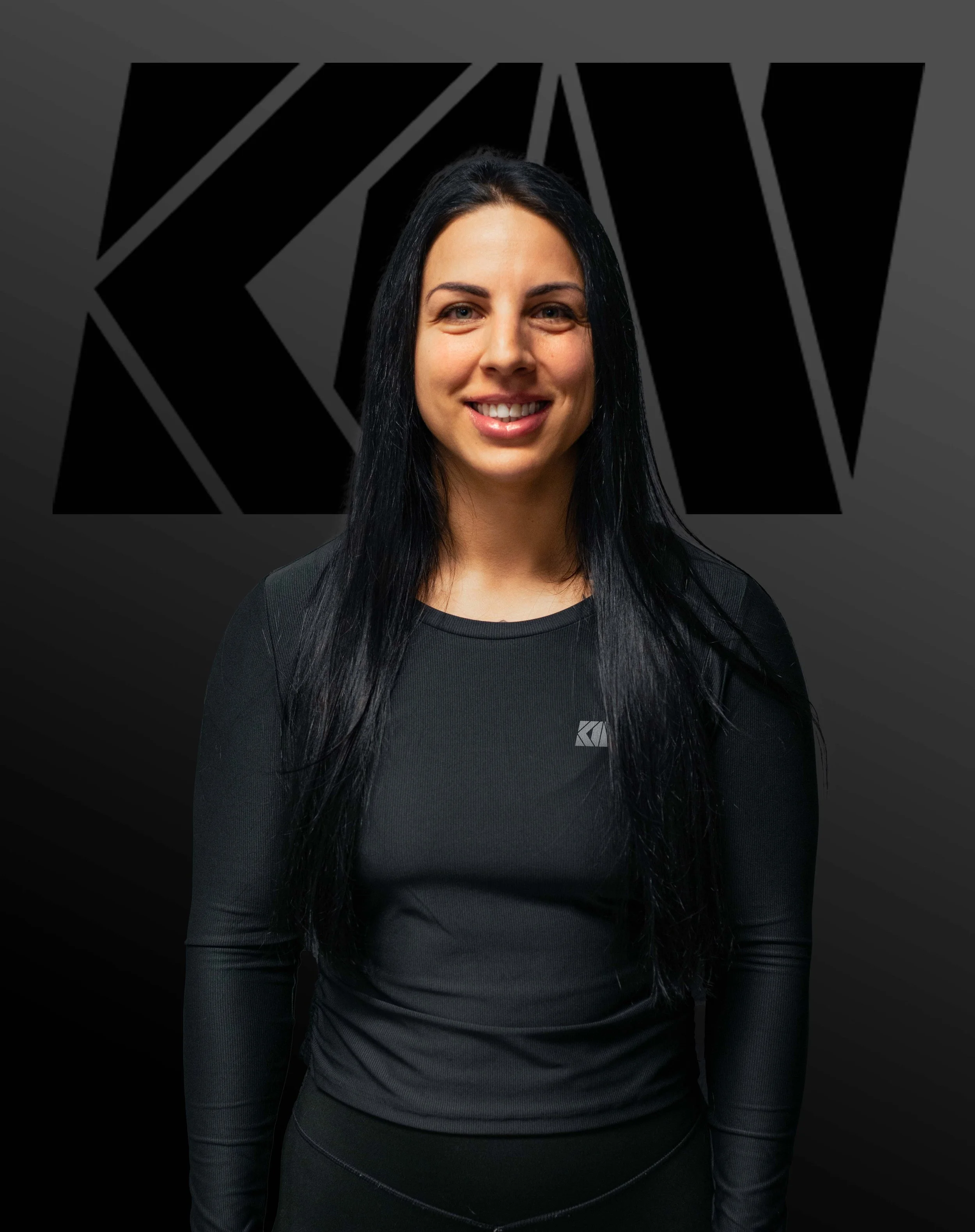 A woman with long black hair smiling, wearing a black long-sleeve shirt with a small logo on the left side, in front of a dark background with a large black geometric design.