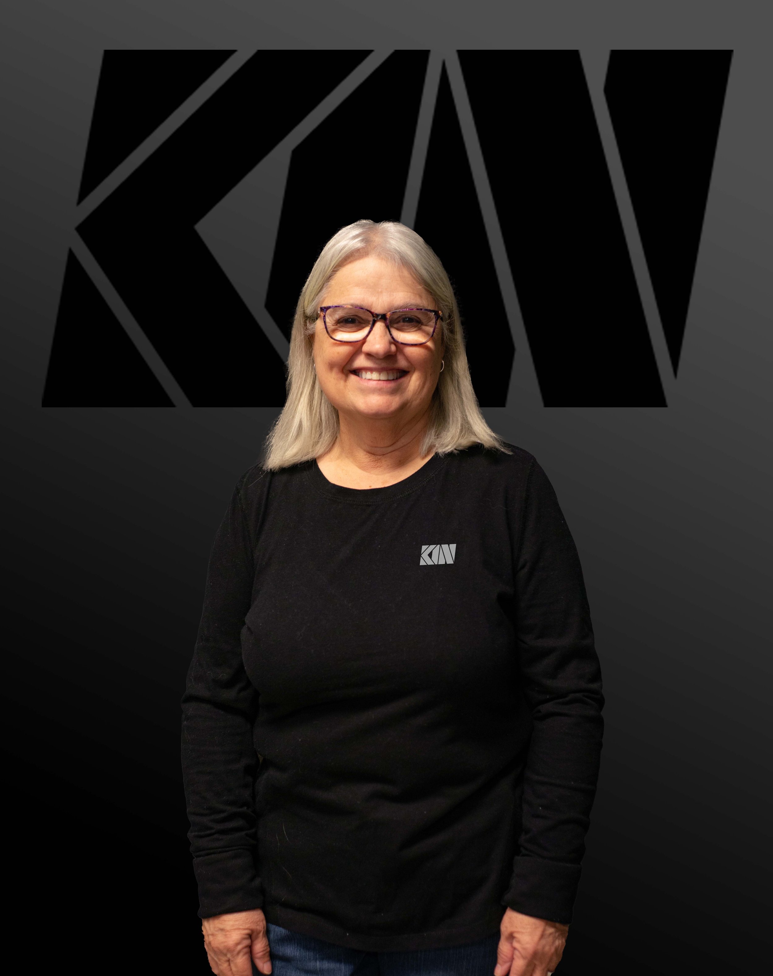 A smiling woman with gray hair, glasses, wearing a black shirt with a logo, standing in front of a dark background with a large black geometric design and the letters 'KAW'.