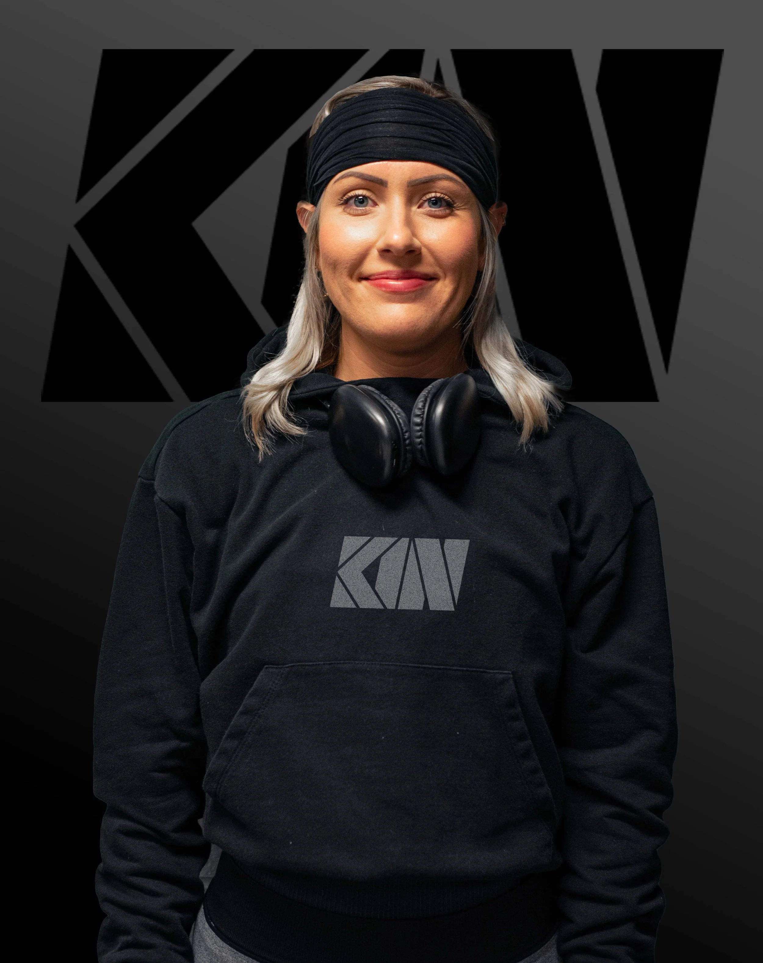 A woman with blonde hair, blue eyes, wearing a black headband, black hoodie, headphones around her neck, and gray pants. She is standing outdoors in front of a dark background with the logo WKTV.