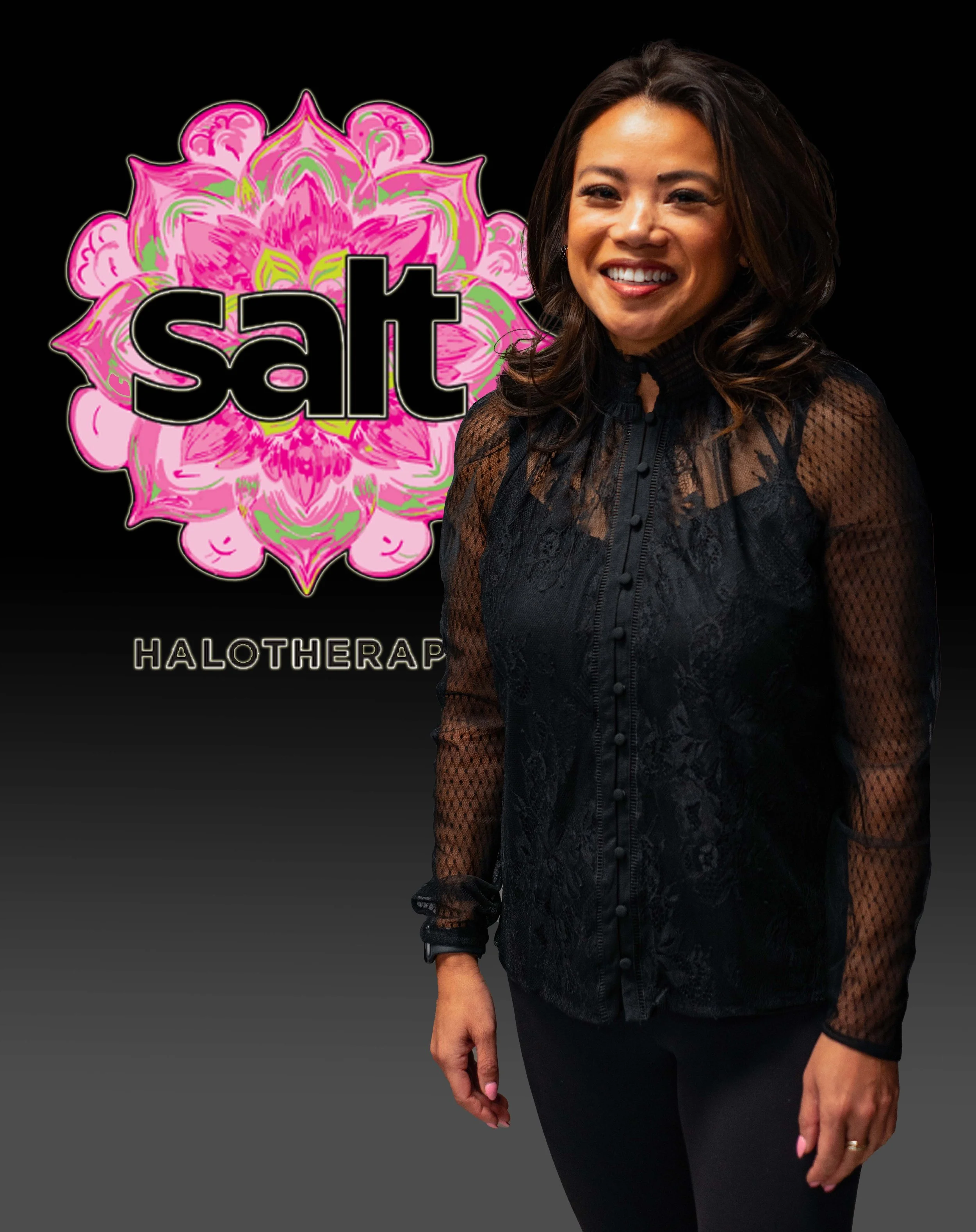 A woman in a black lace blouse smiling, standing in front of a black background with colorful, pink and green floral artwork and the words 'sat HALOTHERAP' visible.