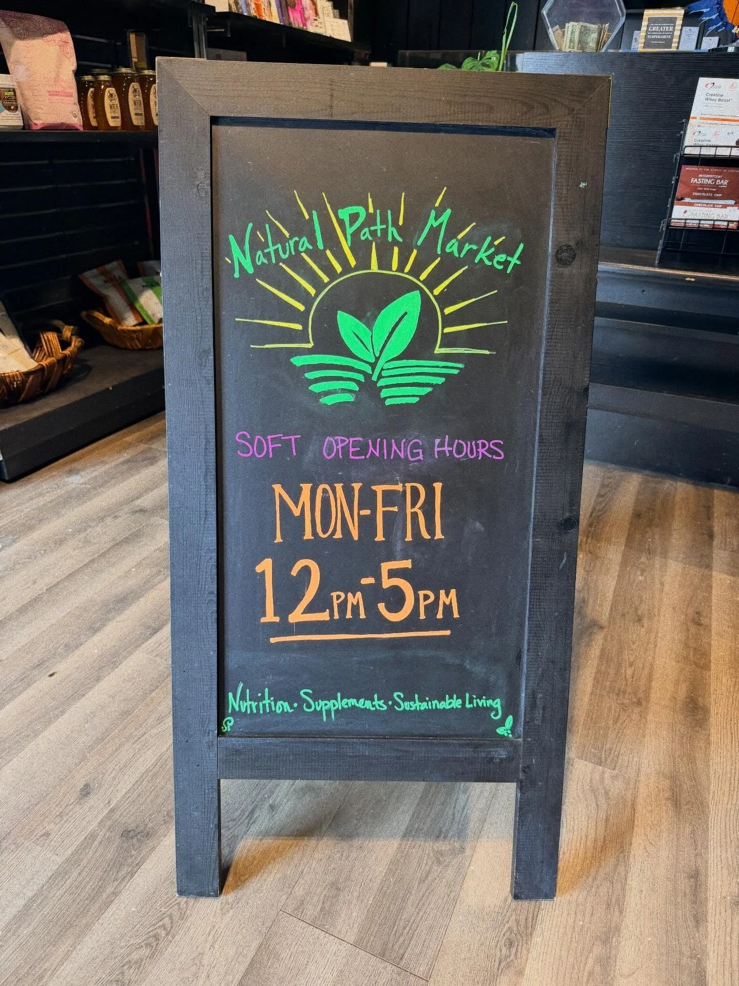 A black chalkboard sign with colorful handwritten text and graphics for a natural market. It displays market hours from 12 PM to 5 PM Monday through Friday, and mentions nutrition supplements and sustainable living.